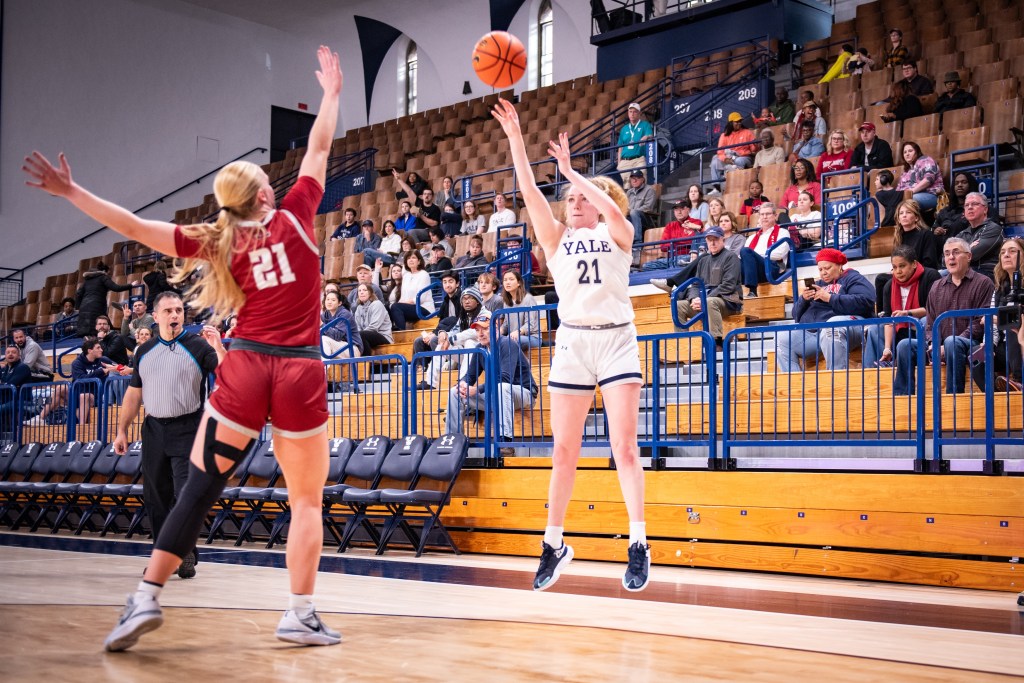 Yale guard Kiley Capstraw follows through as a 3-pointer leaves her hand. She is near the corner of the court, and the crowd in the stands is visible behind her. A Saint Joseph's defender stretches an arm up to try to contest the shot.