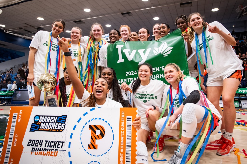 Princeton players pose for a photo with a green Ivy League Tournament championship banner and a commemorative ticket reading, "March Madness, 2024 ticket punched." Many players have streamers around their necks, and forward Chet Nweke sits front and center and punches one fist in the air.