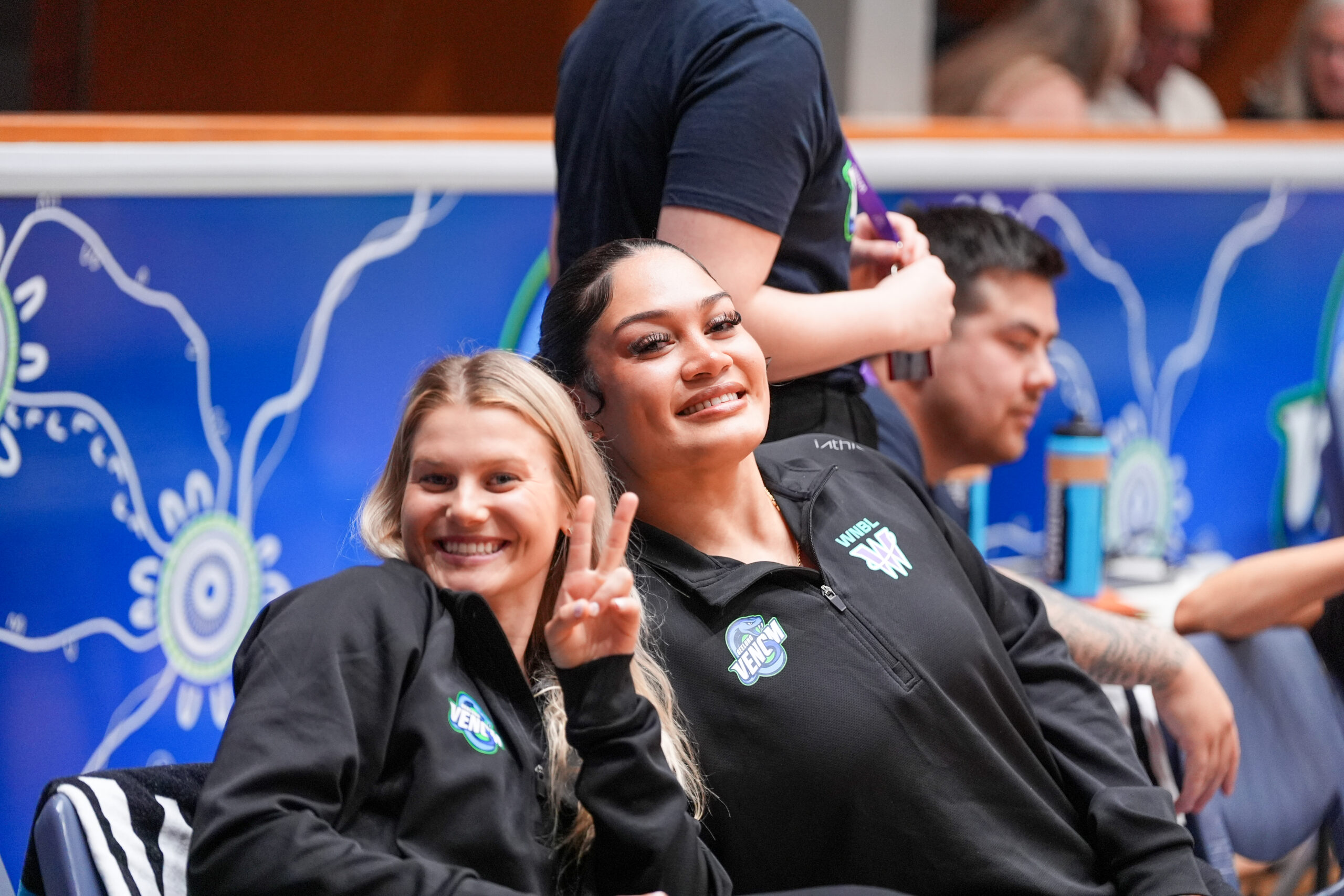 Shyla Heal and Alissa Pili are seated on the bench for the Geelong Venom. They are both leaning into each other and smiling while looking directly at the camera, with Heal gesturing a peace symbol with her fingers.