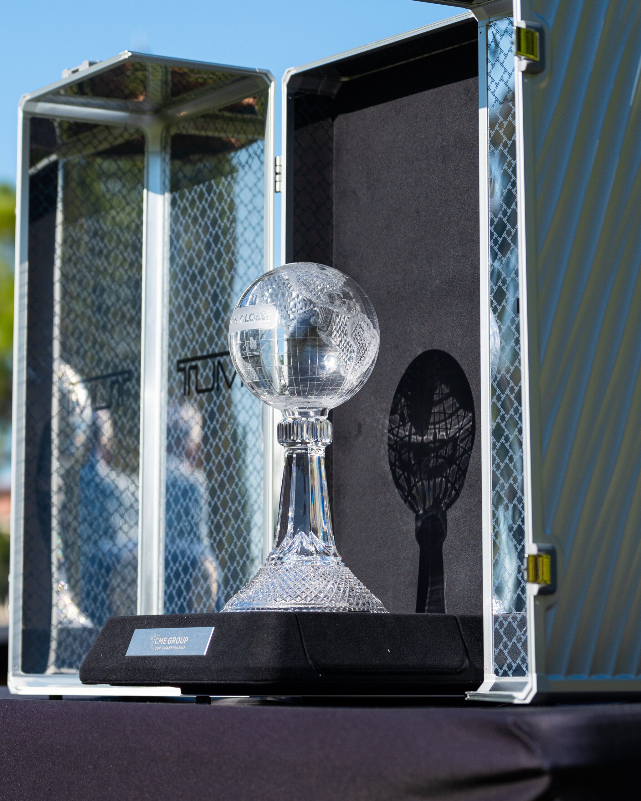 A crystal globe trophy for the CME Group Tour Championship sits atop a black trophy stand. The TUMI travel case surrounds the trophy. The sun shines on the crystal globe, making the trophy shine and casting a shadow on the black travel case background.