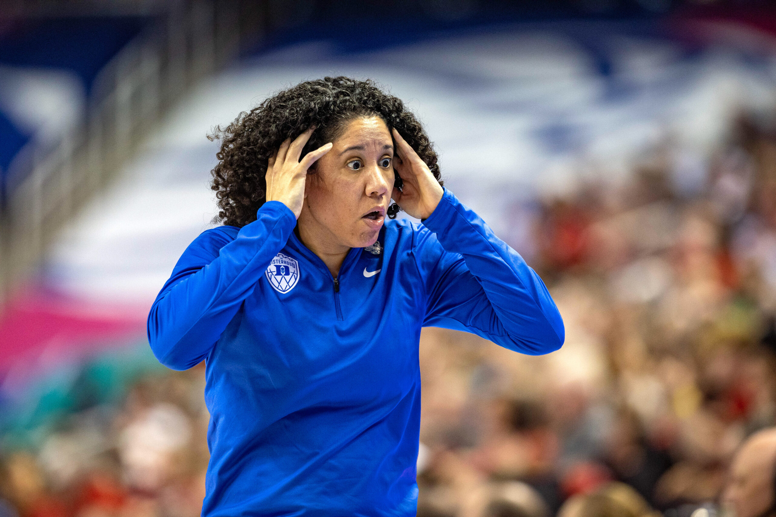 Duke head coach Kara Lawson holds her temples with spread fingers while looking off-screen with a bewildered expression. A packed section of out-of-focus fans is behind her in the background, with a section adjacent to them covered by an equally out-of-focus ACC logo.