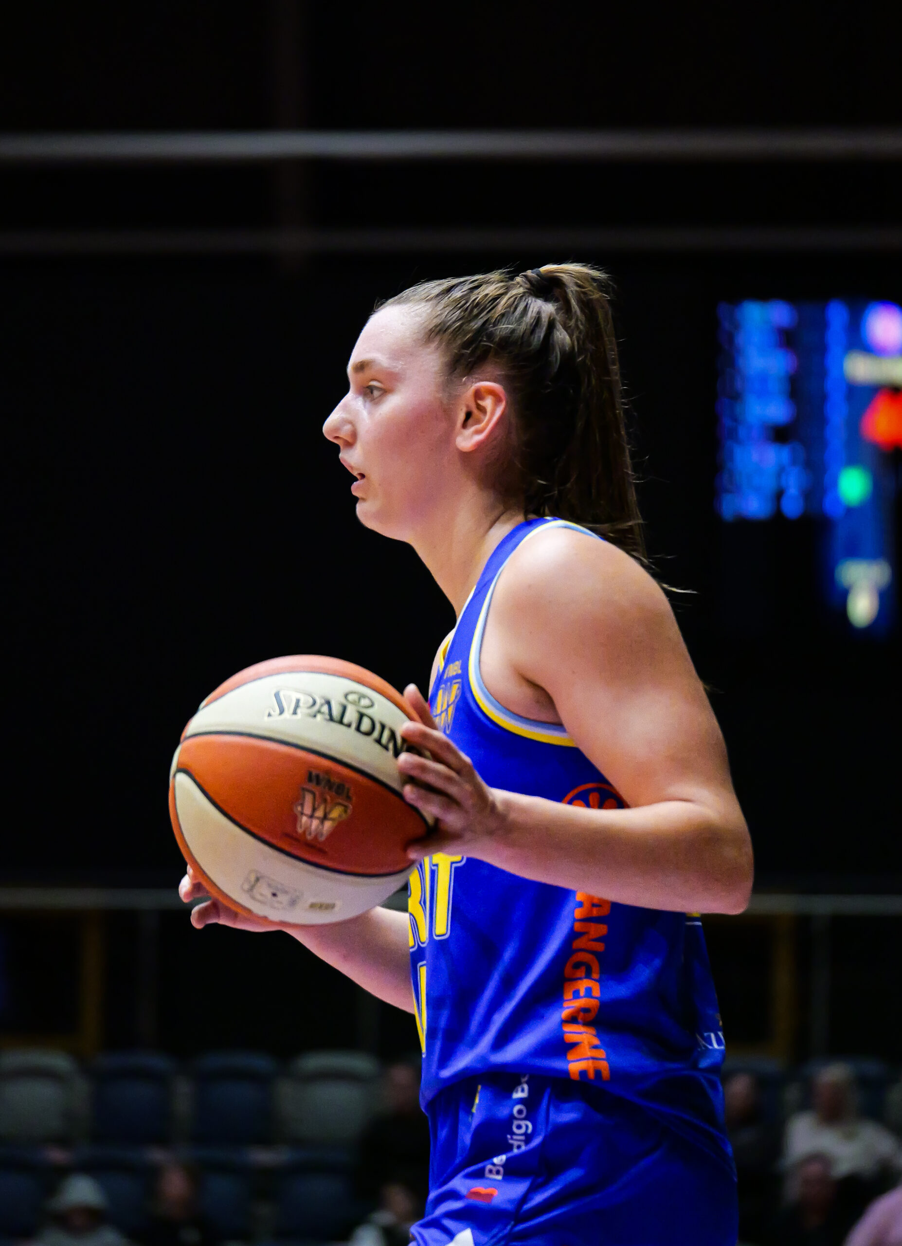 A side profile of Isobel Borlase caressing a basketball at the top of its dribble as she brings the ball up the court for the Bendigo Spirit. Half of the scoreboard is visible in the background of the image but the details are completely blurry.