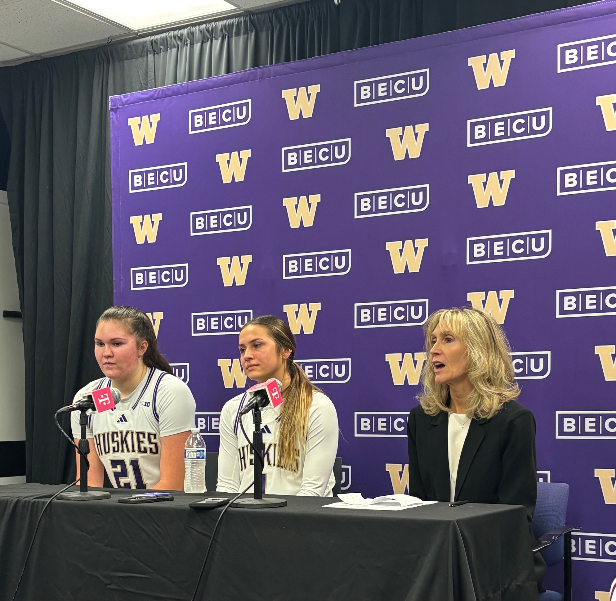 Seated at a table with a black tablecloth and two microphones, from left to right, are University of Washington forward Brynn McGaughy, #21, and guard Hannah Stines, #1, wearing their white home basketball jerseys, as well as Head Coach Tina Tangley. Behind the trio is a purple back drop with gold University of Washington logos.
