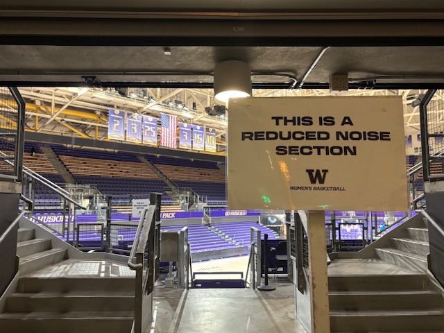 A white sign with the words "this is a reduced noise section" and the University of Washington Women's Basketball logo written in purple is posted outside one of the sections of the arena with the edge of the basketball court and bleachers seen in the background.