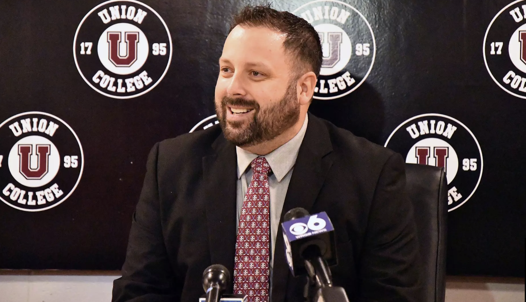 Union College women's hockey coach Tony Maci stands at a podium in front of several microphones.
