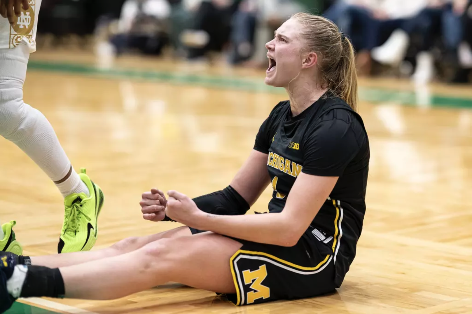 Michigan women's basketball guard Olivia Olson sitting on the floor yelling, wearing blue jersey with maize trim.