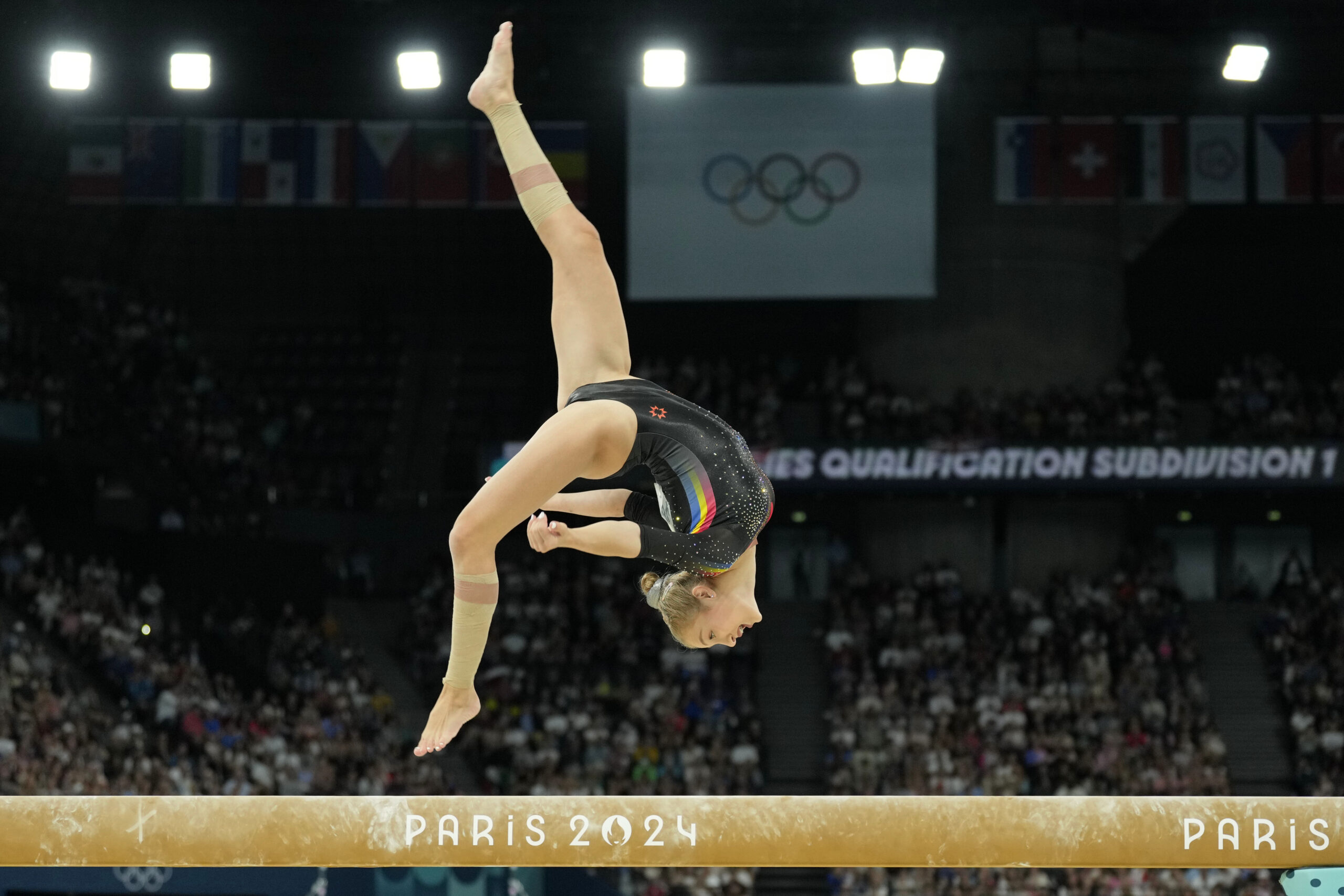 An athlete in a black leotard performing a back layout stepout on the balance beam.