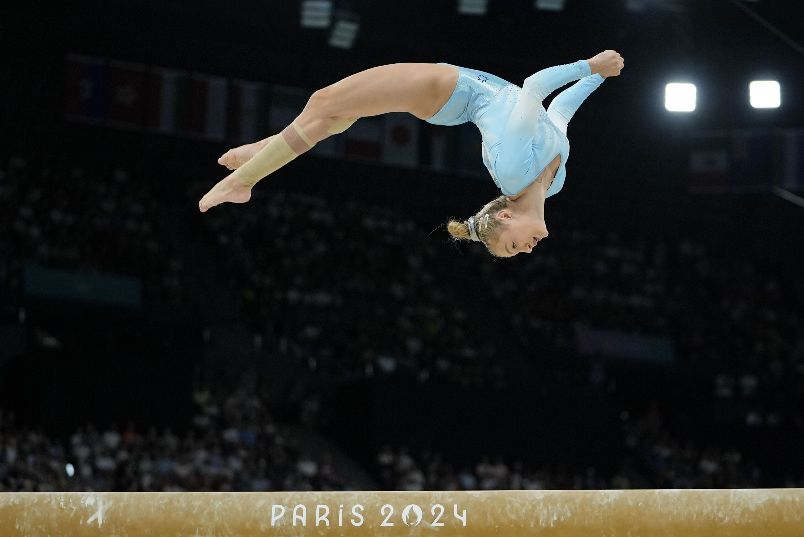 A gymnast in a light blue leotard does an arching back flip over the balance beam.
