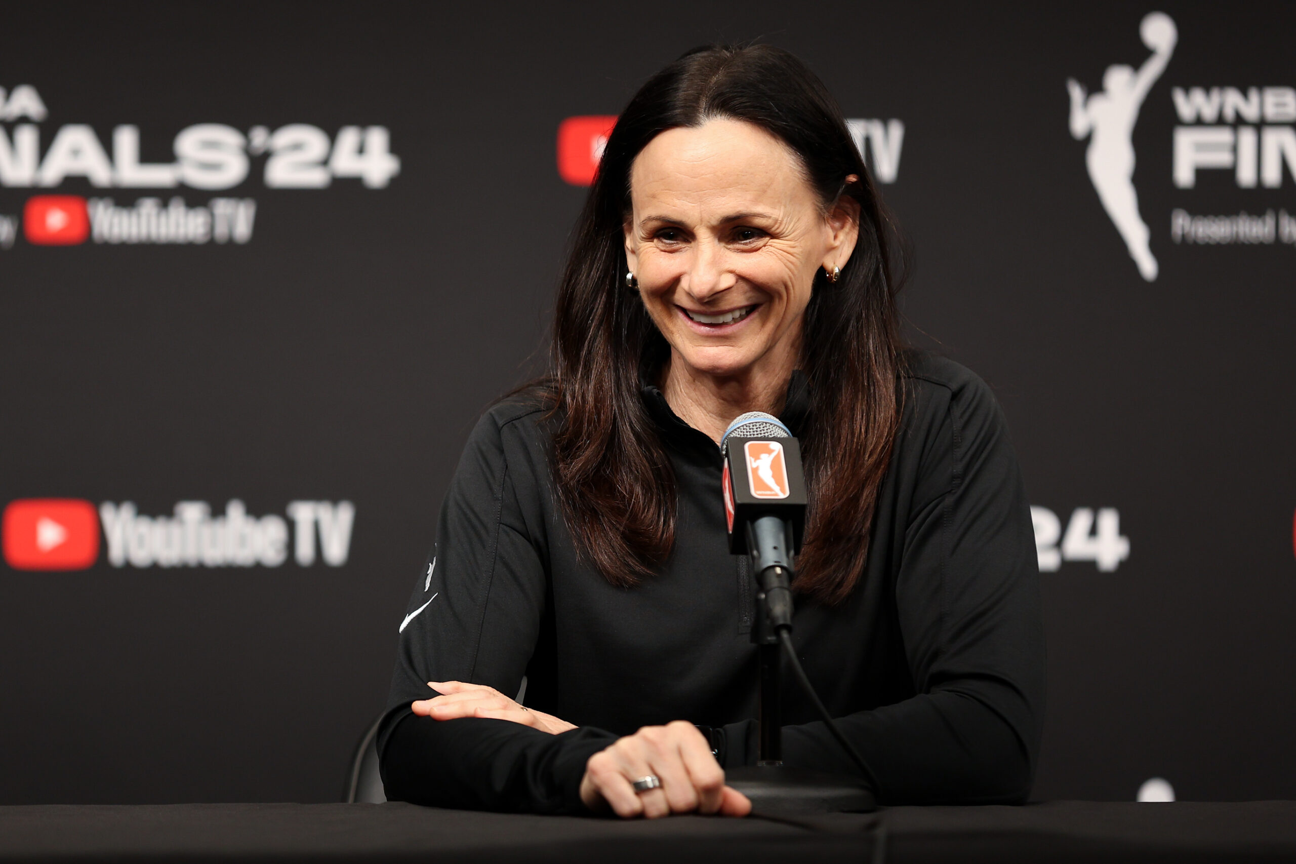 Sandy Brondello sits in front of a WNBA microphone smiling. The backdrop with has the 2024 WNBA Finals logo on it is blurred in the background.