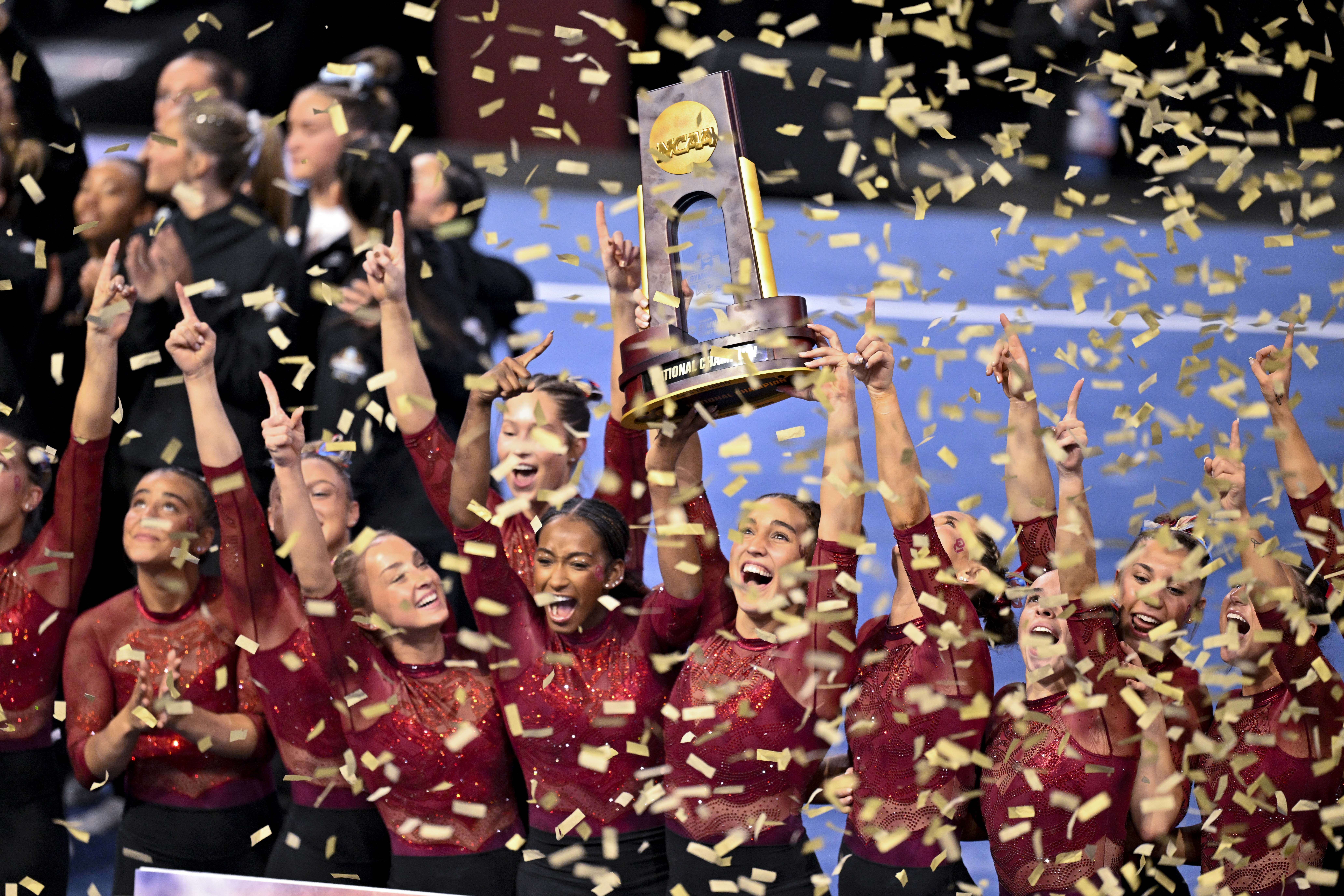 A group of female gymnastics in dark red leotards celebrates a trophy as confetti falls around them at the 2025 NCAA gymnastics championships.