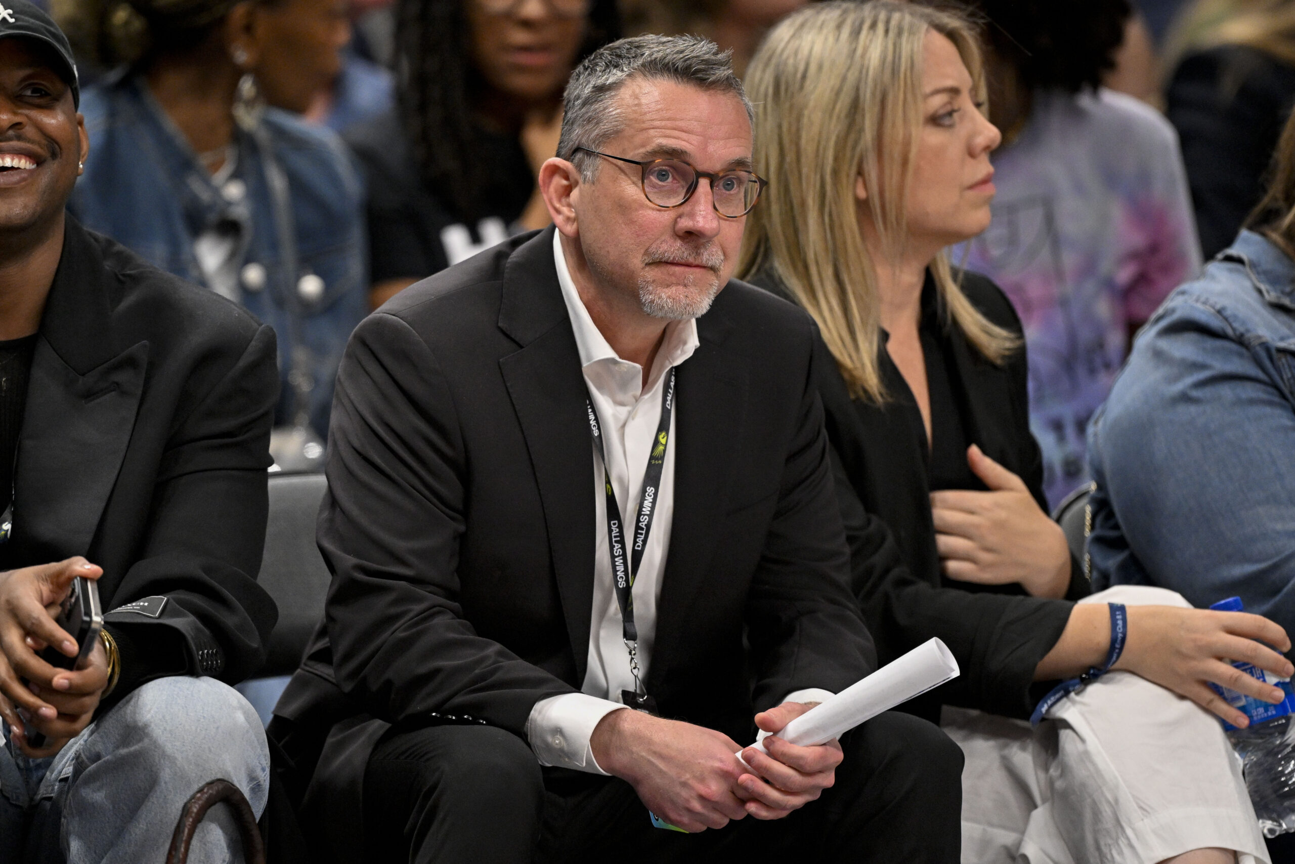 Dallas Wings general manager Curt Miller is sitting down in a chair. He has a neutral expression while holding a rolled up paper in his hands.