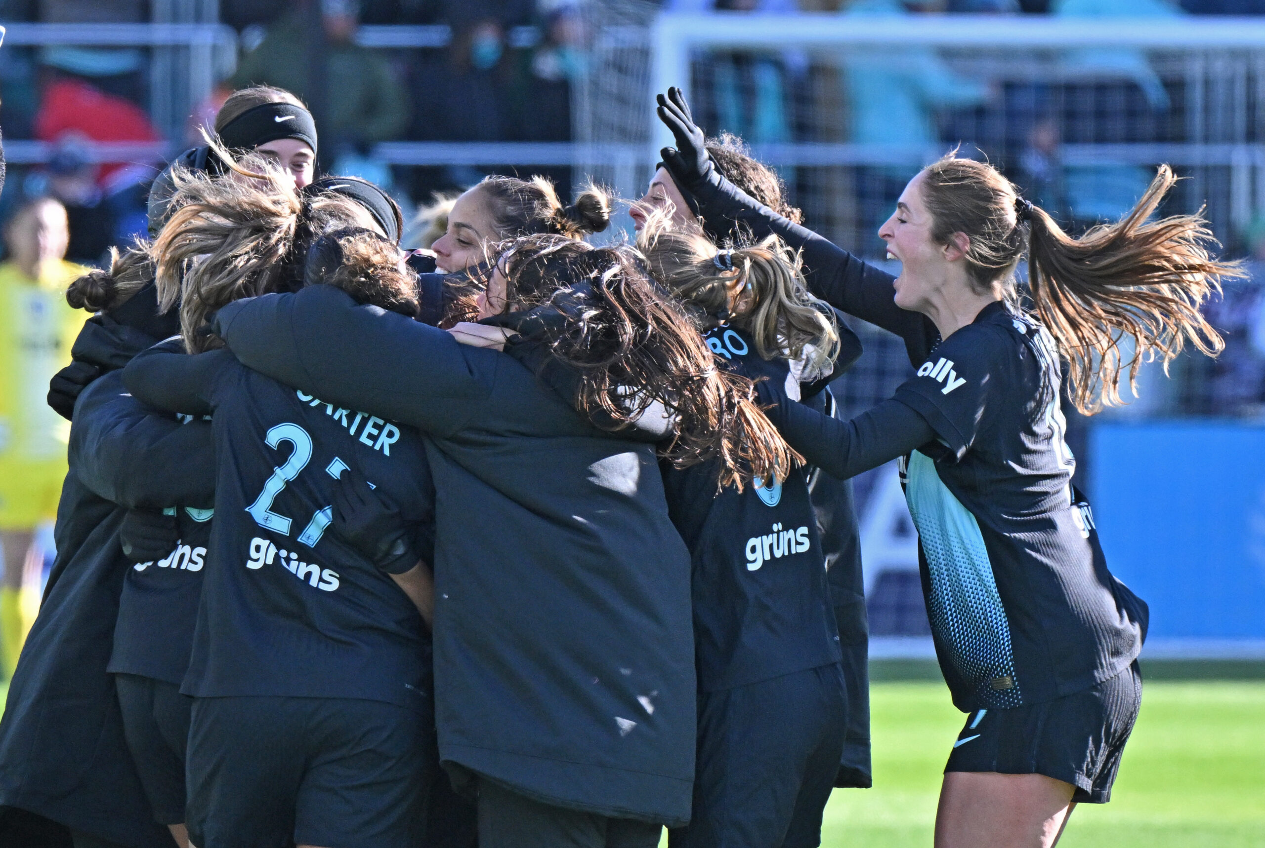 Gotham FC players celebrate on the pitch. They are all standing in a huddle, smiling and cheering.