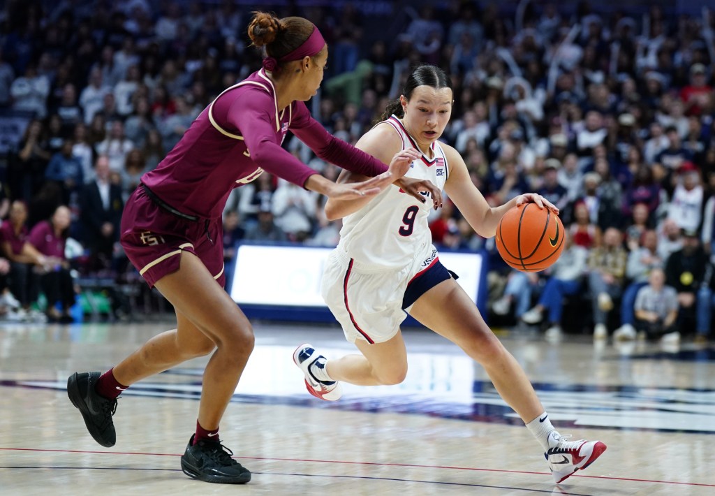 UConn guard Kayleigh Heckel drives to the basket with her left hand while facing defensive pressure from Florida State guard Tatum Greene. Greene runs alongside Heckel and tries to keep her at bay with her left arm.