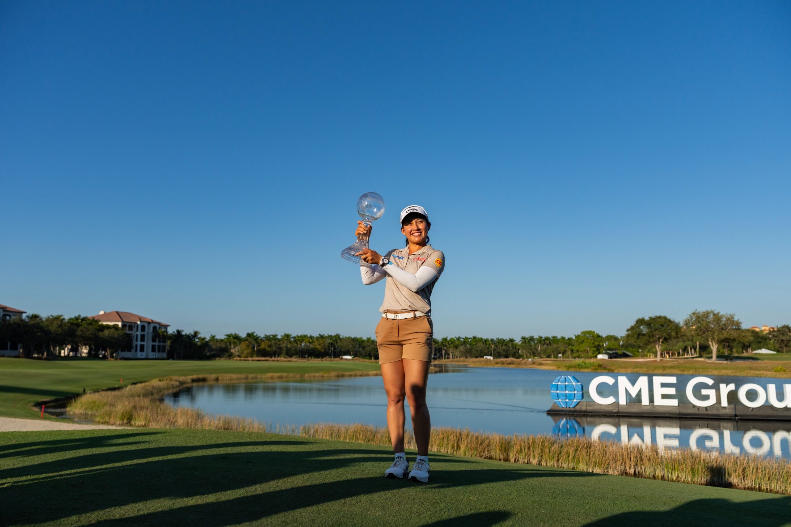 Jeeno Thitikul, winner of the 2025 CME Group Tour Championship, poses with the crystal globe trophy and holds it up to her right. The CME Group sign is on the water of the pond in the background. The golf course spreads behind her, and she stands on the green of the 18th hole.