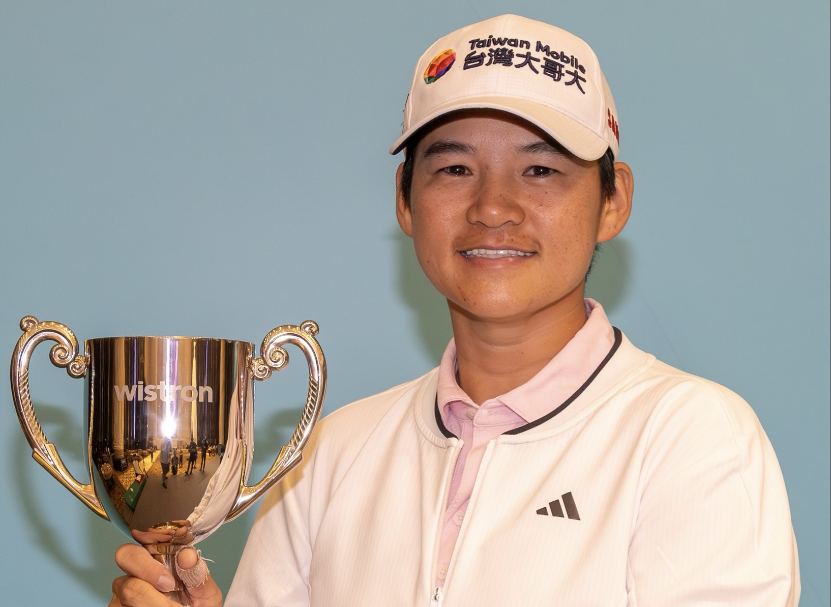 Yani Tseng, winner of the 2025 Wistron Ladies Open, holds the gold tournament trophy up to her right in front of a light blue background. She wears her golf tournament clothes and hat, and she smiles at the camera.