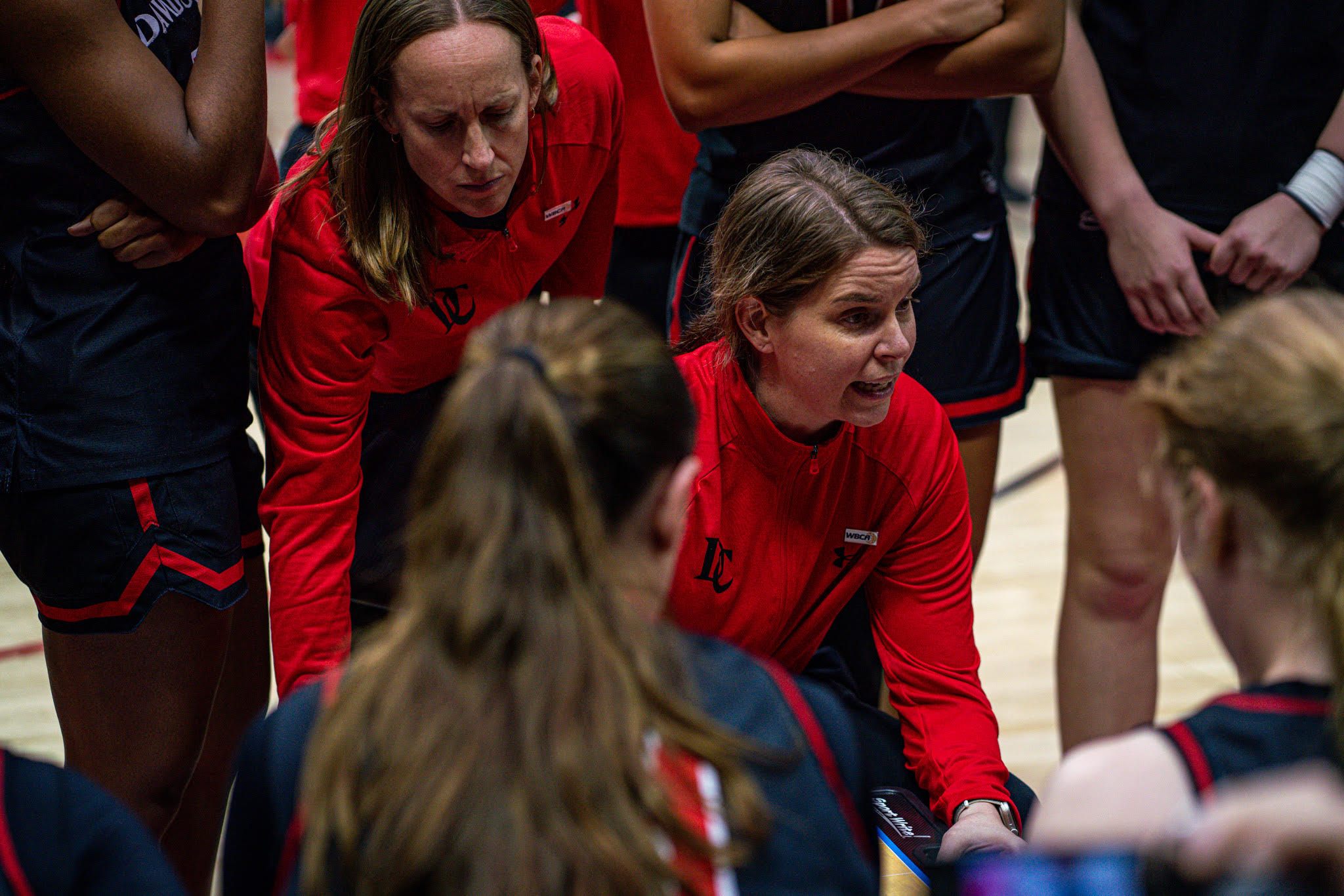 Davidson head coach Gayle Fulks sits in the middle of her players, who are in a circle around her, during a timeout. A member of her staff is leaning over behind her.