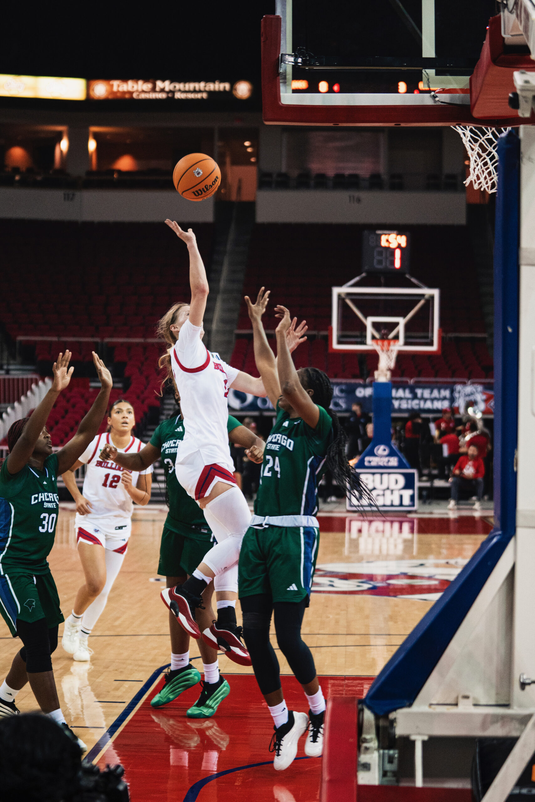A player jumps up to make a basket. She is wearing a white uniform and she is being guarded by a player in a green uniform.