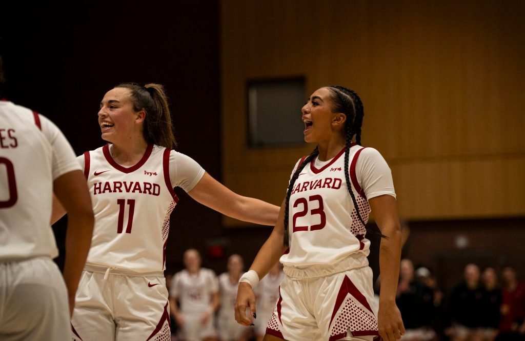 Sophomore guards Alayna Rocco and Nina Emnace smile and look at something to their right outside the frame. Both are wearing white Harvard jerseys with crimson trim and lettering.