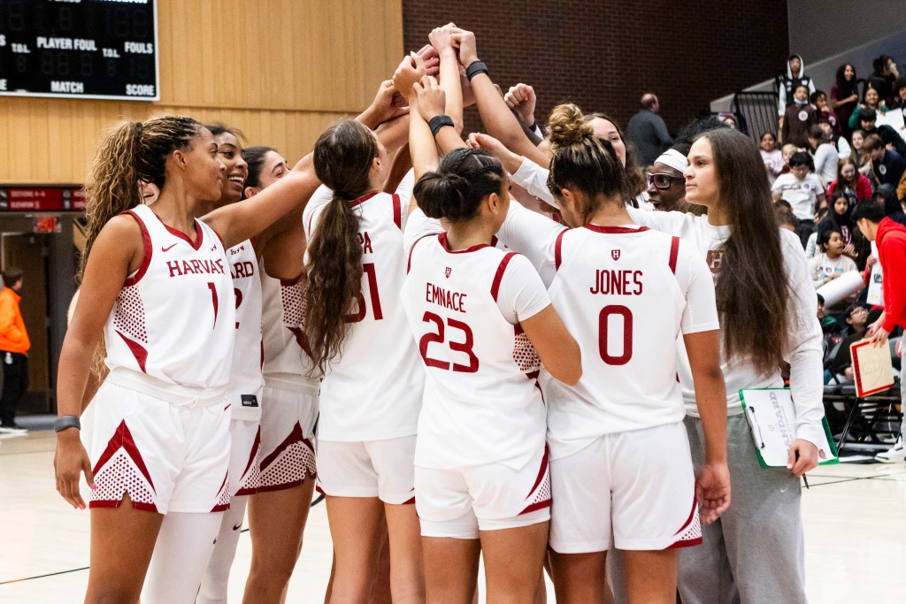 Harvard players huddle tightly on the court. They each raise an arm toward the center of the huddle. Children from local schools are visible in the stands behind them.