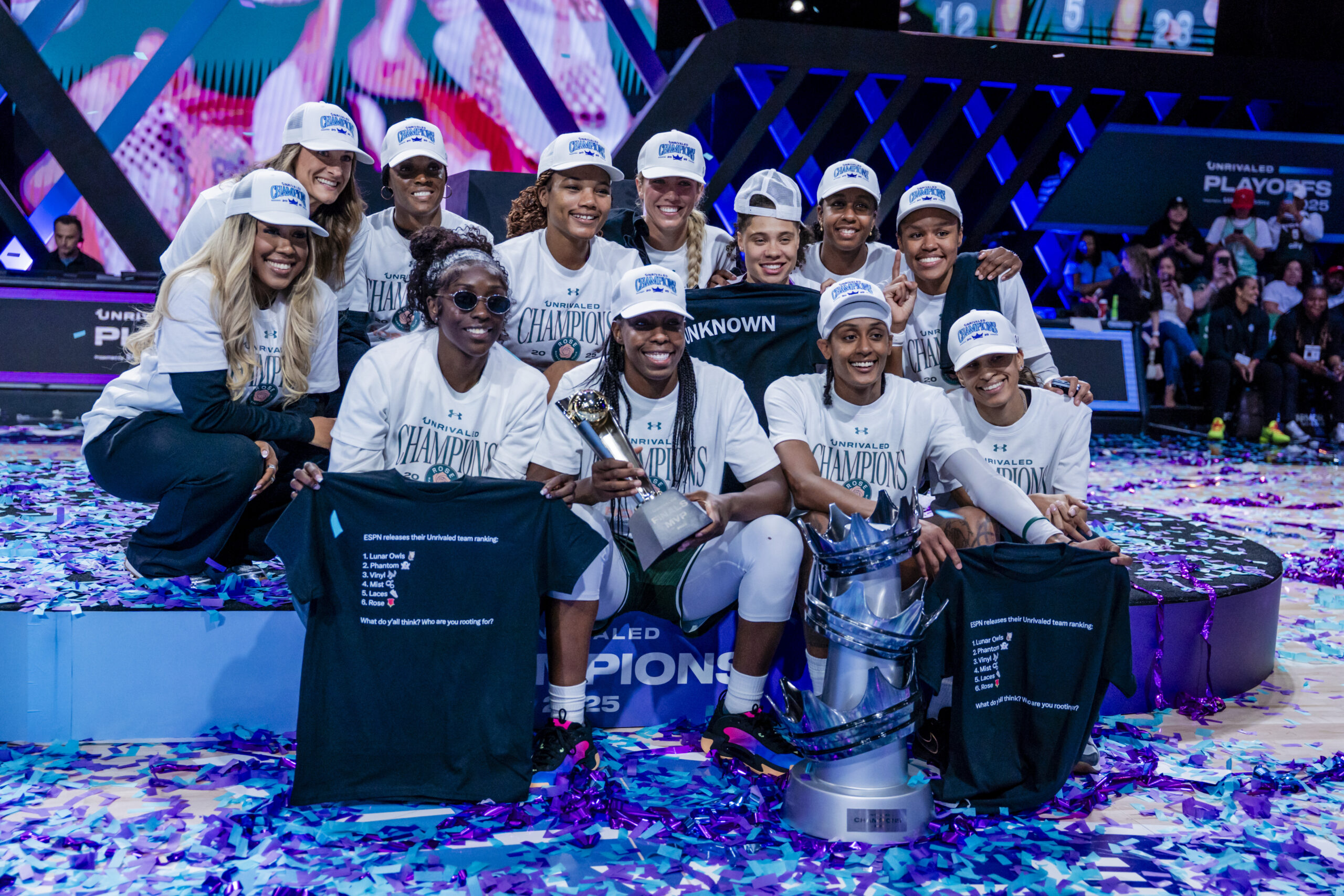 Rose BC teams gather to take a photo with smiles on their faces. They are sitting on a podium and the trophy is on the ground in front of them with confetti spreading everywhere.