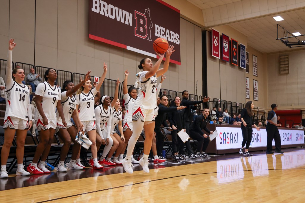 Brown point guard Grace Arnolie shoots an open 3-pointer in front of the Brown bench. The bench players are standing, and many of them are making 3-point signs with their hands in anticipation.