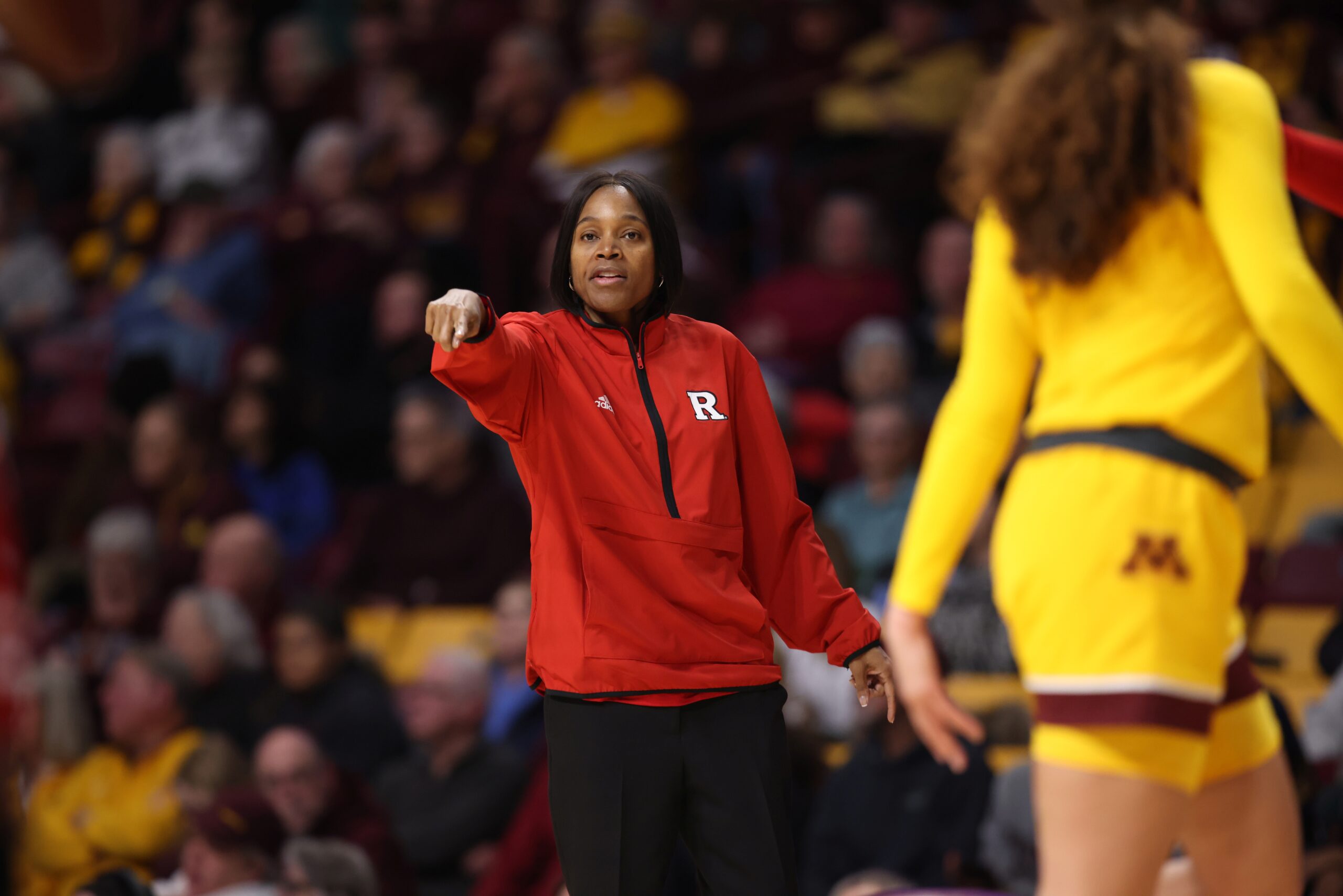Rutgers head coach Coquese Washington points out and down while looking toward the camera, as a Minneosta Gophers player walks by. A packed crowd is sitting out-of-focus in behind Washington.