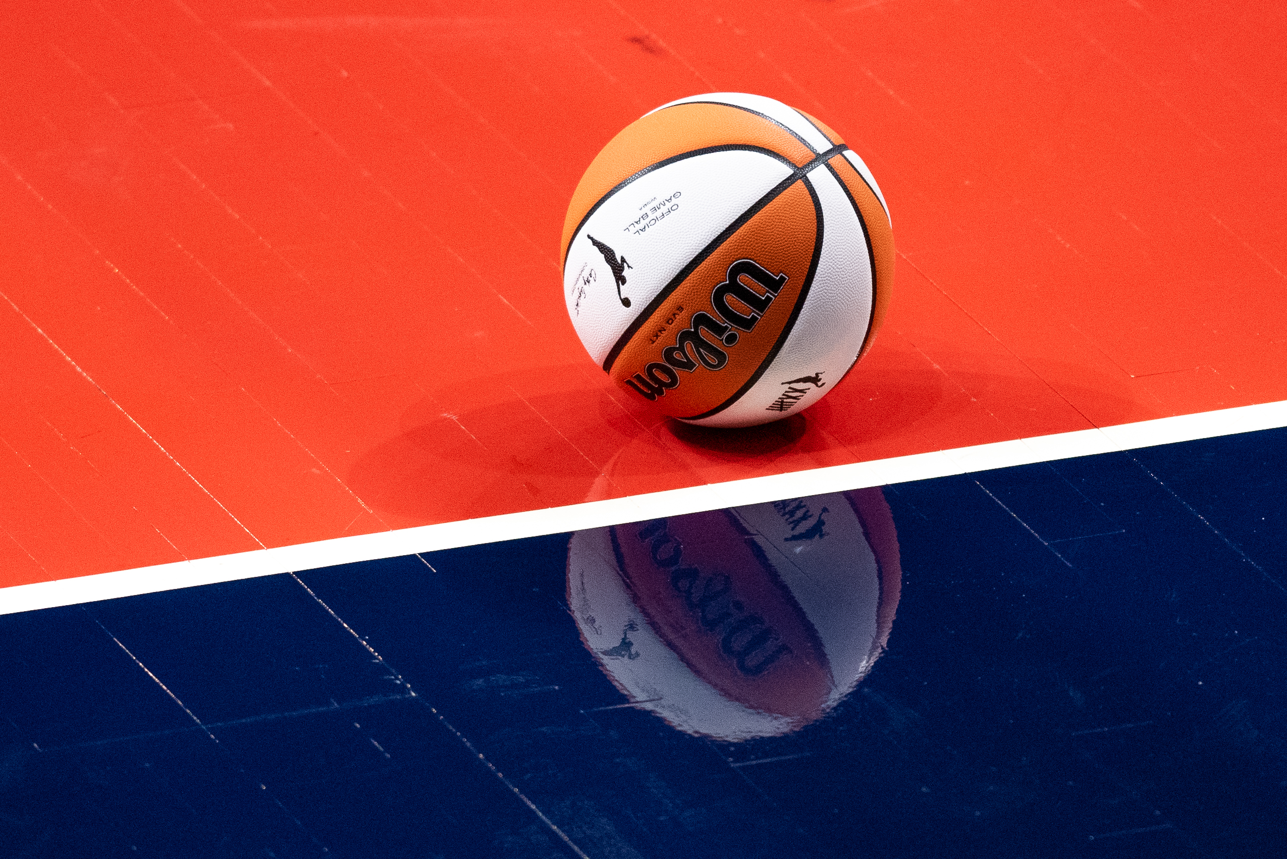 An orange and white banded WNBA basketball sits on a red court, with a white stripe on the court leading into blue court in the foreground