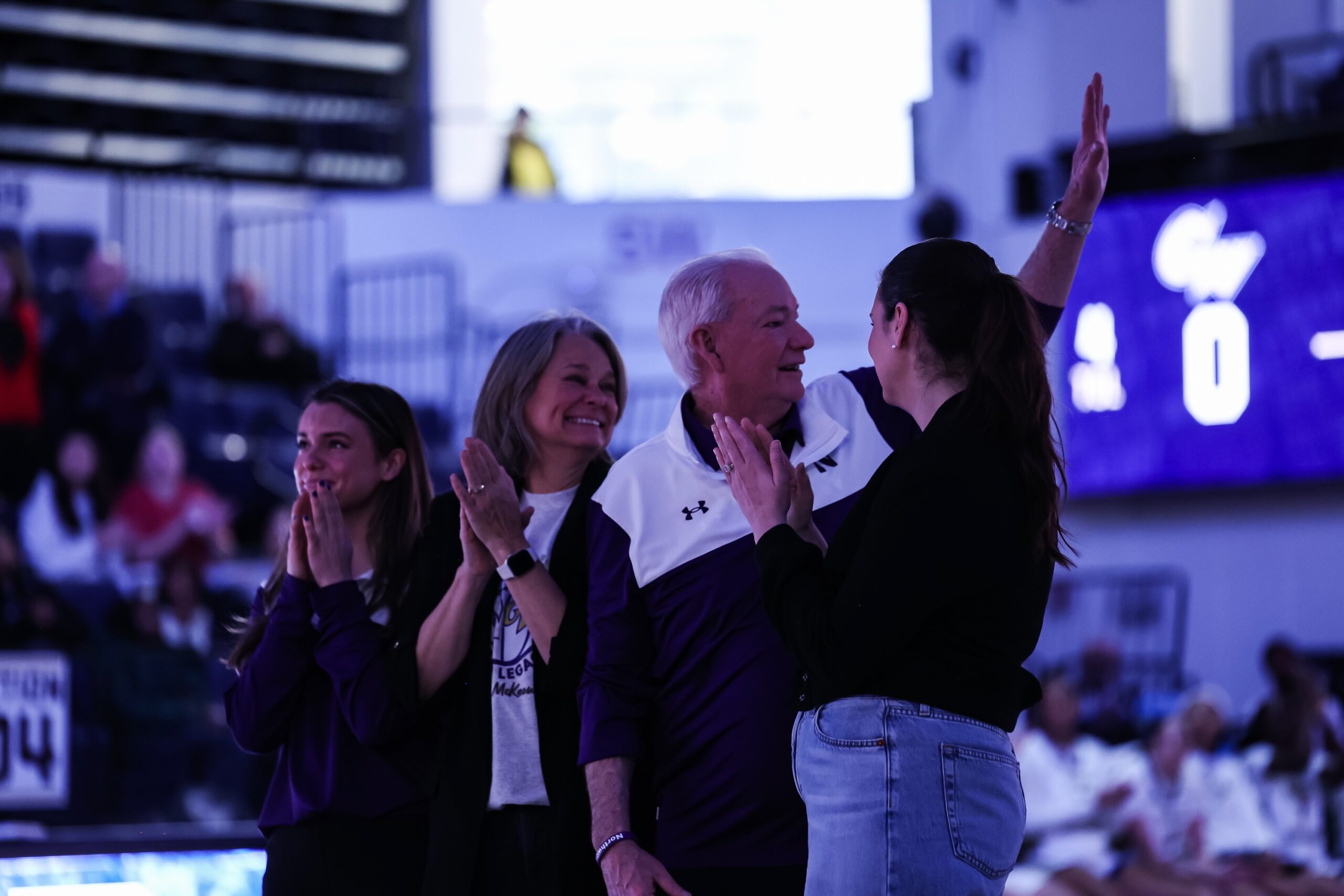 Northwestern head coach Joe McKeown is standing with his wife and two daughters during a pregame ceremony. He turns over his left shoulder and waves to the crowd with his left hand as his family claps.