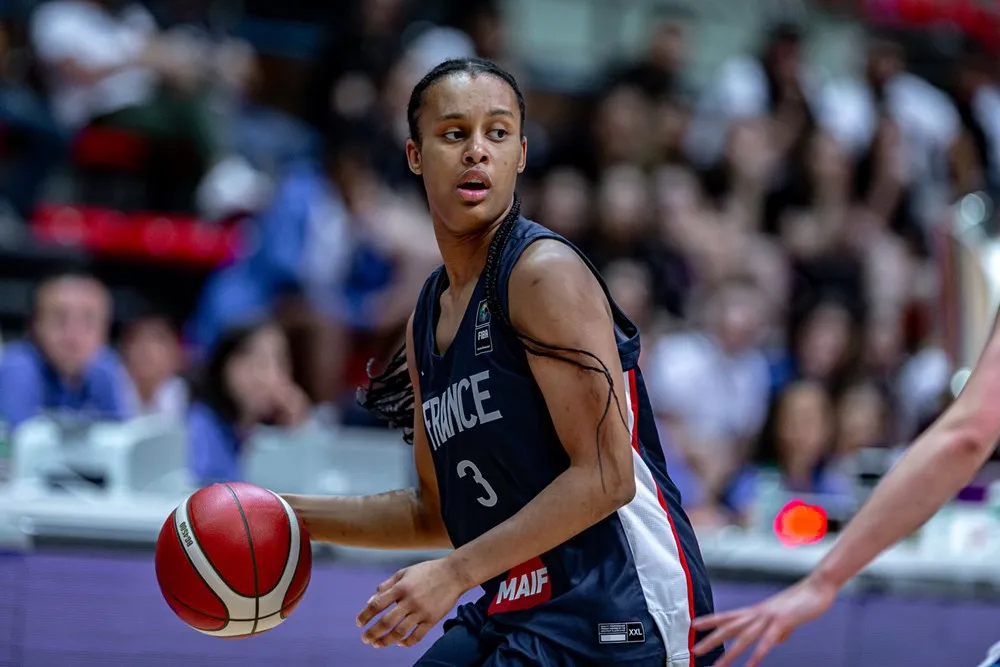 French basketball player Nell Angloma dribbles the ball while looking off-screen toward the rest of the court. Fans sit out-of-focus in the background behind her.