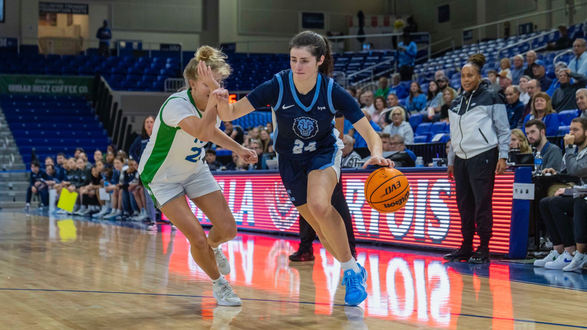 Columbia guard Riley Weiss dribbles the ball with her left hand and uses her right arm to protect the ball from an FGCU defender. She is right on the 3-point line and in front of the FGCU bench.