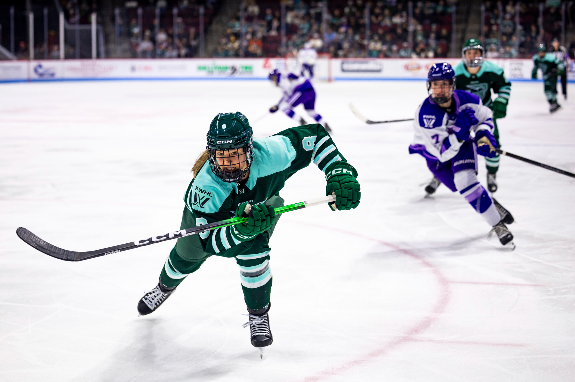 Winn leans forward, holding her stick in her hands as she hunts down the puck, which is out of frame in the corner. She is wearing a green home uniform.