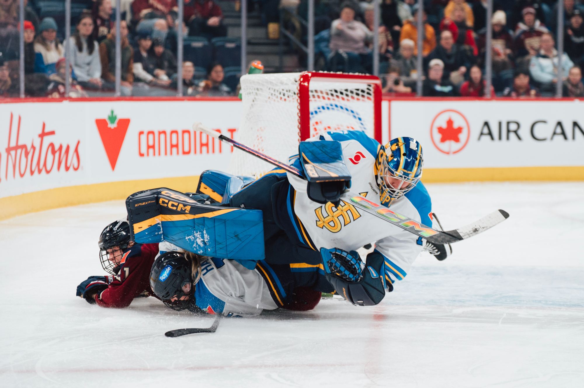 A Montrรฉal and Toronto player are underneath a nearly horizontal Kirk as she attempts to make a save just outside her crease. The Montrรฉal player is in a maroon home uniform, while Kirk is in a white away one.