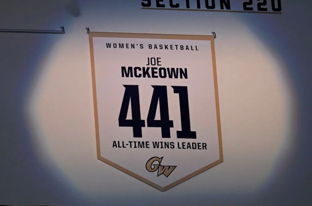 A spotlight shines on a white banner with gold trim. The banner is shaped like a square but with the bottom pointing down. The banner prominently displays the number 441 and reads in all caps, "Women's basketball, Joe McKeown, all-time wins leader."