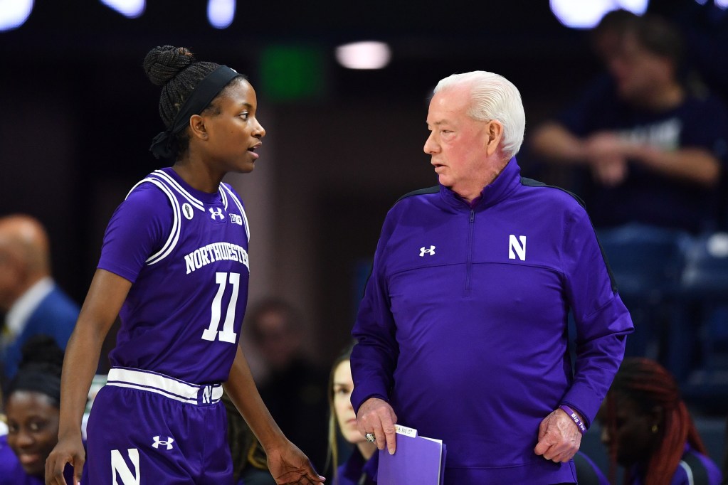 Northwestern head coach Joe McKeown and guard Hailey Weaver stand next to each other and talk during a game. Waver is wearing a bright purple jersey, and McKeown is wearing a quarter-zip in the same shade.