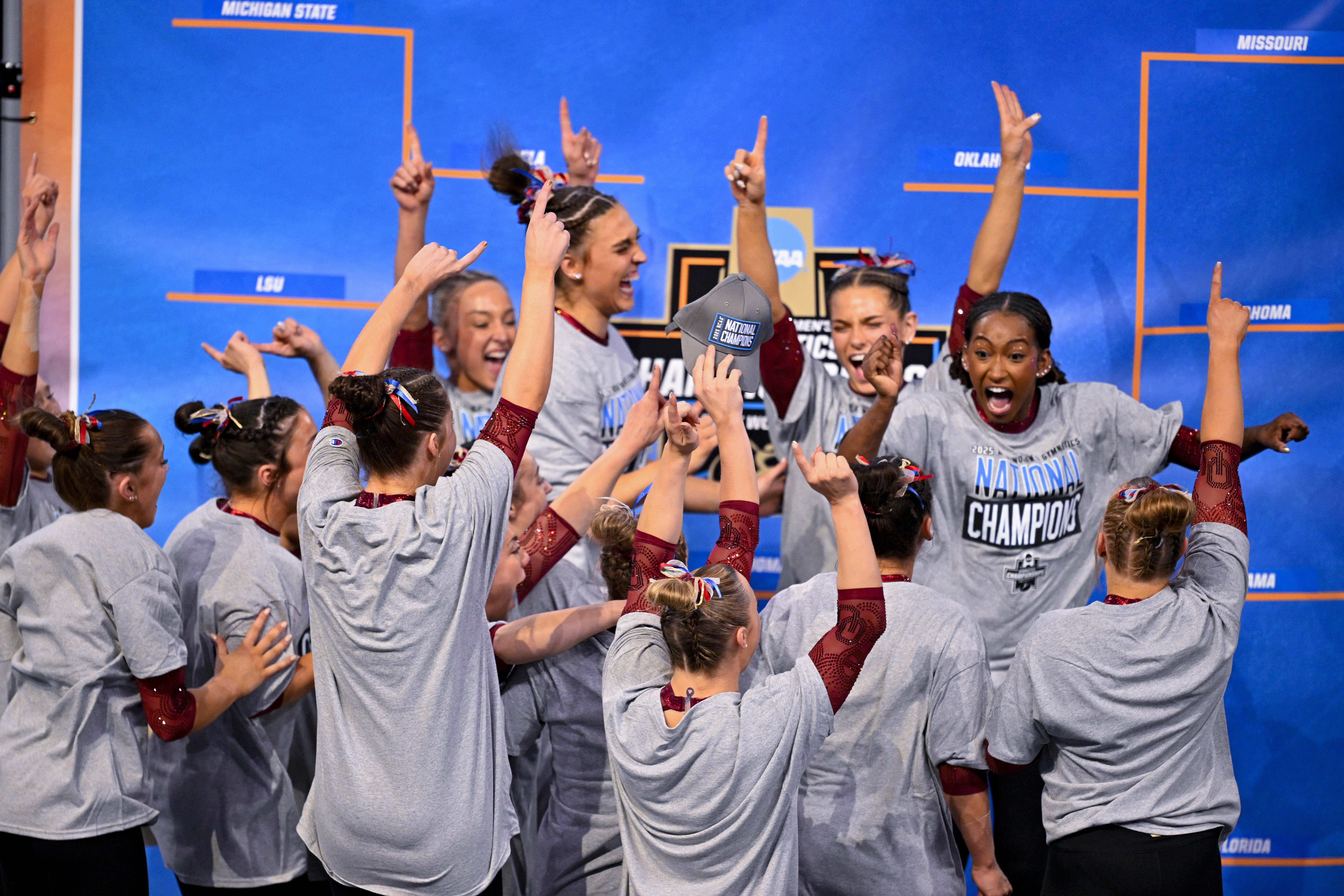 Members of the OU women's gymnastics team, dressed in gray t-shirts, wave the number 1 in the air after winning NCAAs in 2025.