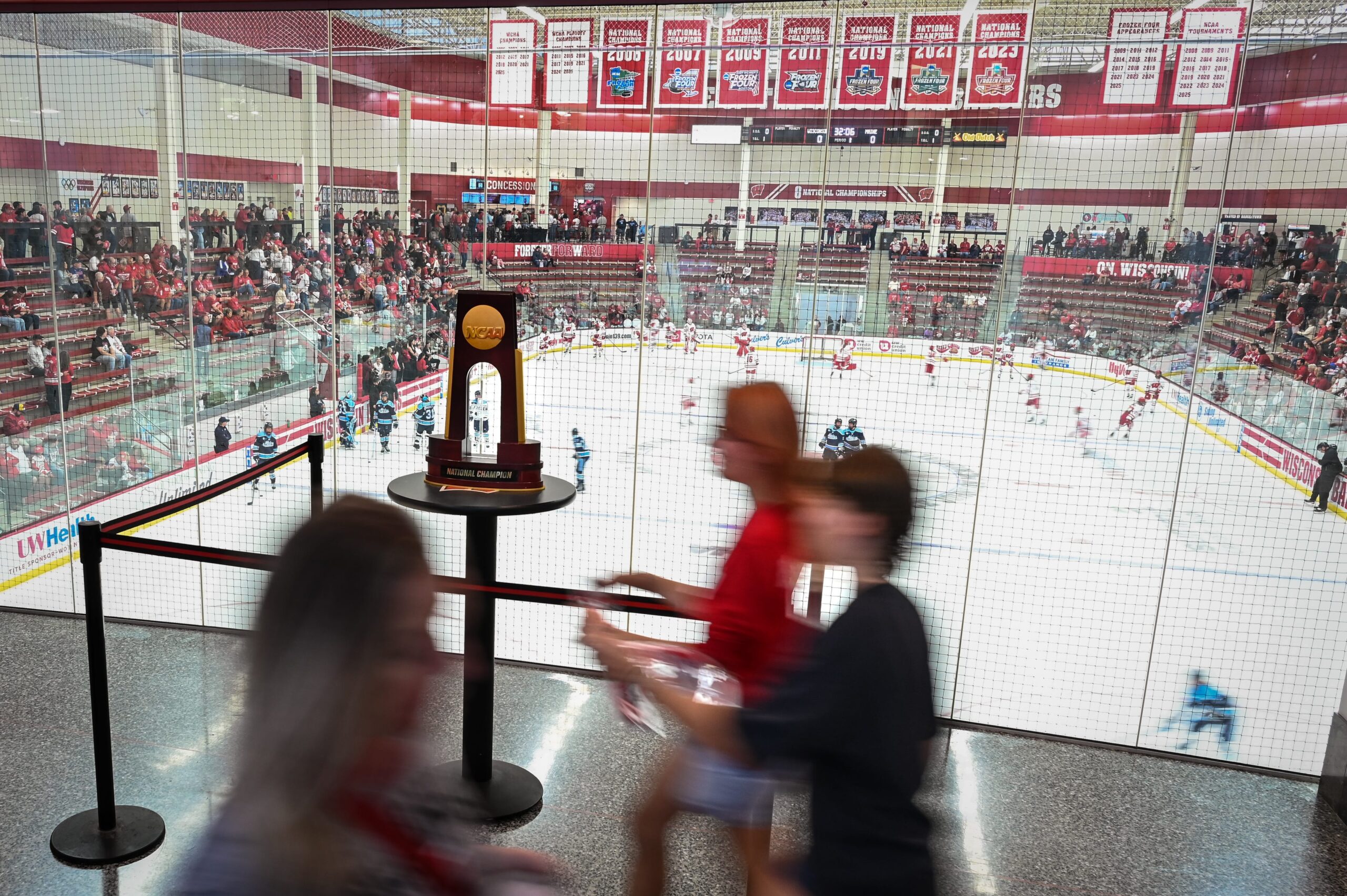 Fans walk past the 2025 NCAA women’s hockey national championship trophy which is placed on a small high-top table in Wisconsin's home hockey arena