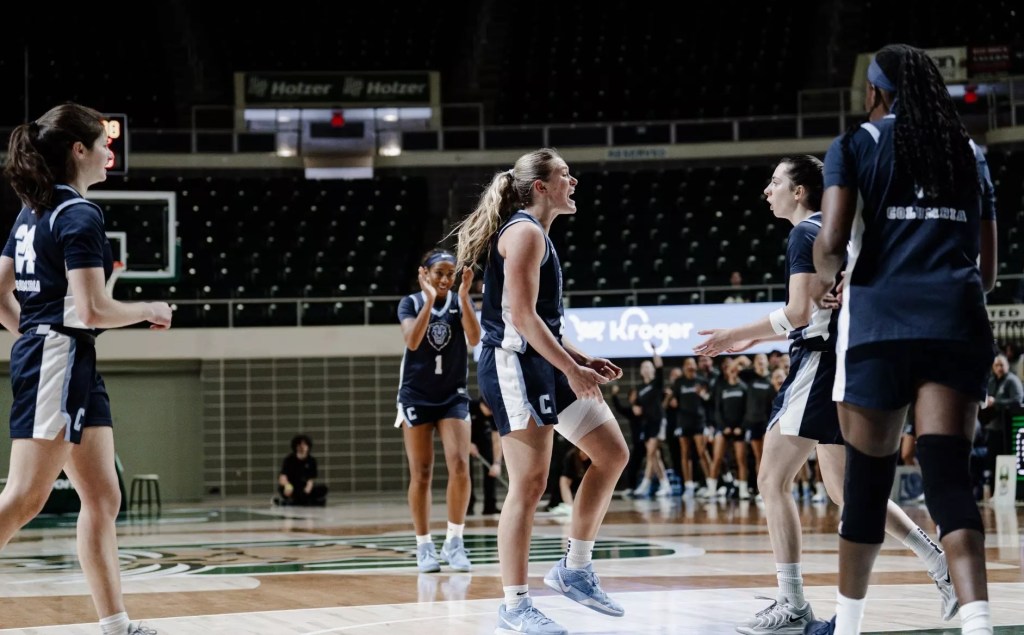 Columbia guard Fliss Henderson is shown from the side, seemingly shouting in celebration as she approaches guard Marija Avlijas. Three other teammates walk toward them from different directions, and one of them claps her hands and smiles.