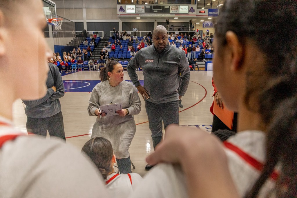 American head coach Kelly Killion holds a whiteboard and a marker. She sits in front of her players, who are on the bench, during a timeout. Associate head coach Robert Isme stands next to her with one hand on his hip.