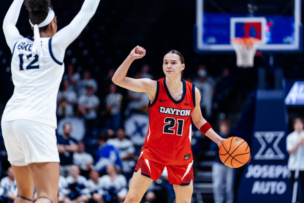 With a defender in front of her, Dayton graduate student guard Nicole Stephens makes a play call by raising her right fist to head level while her upper arm is parallel to the ground. She holds the ball in her left palm mid-dribble.