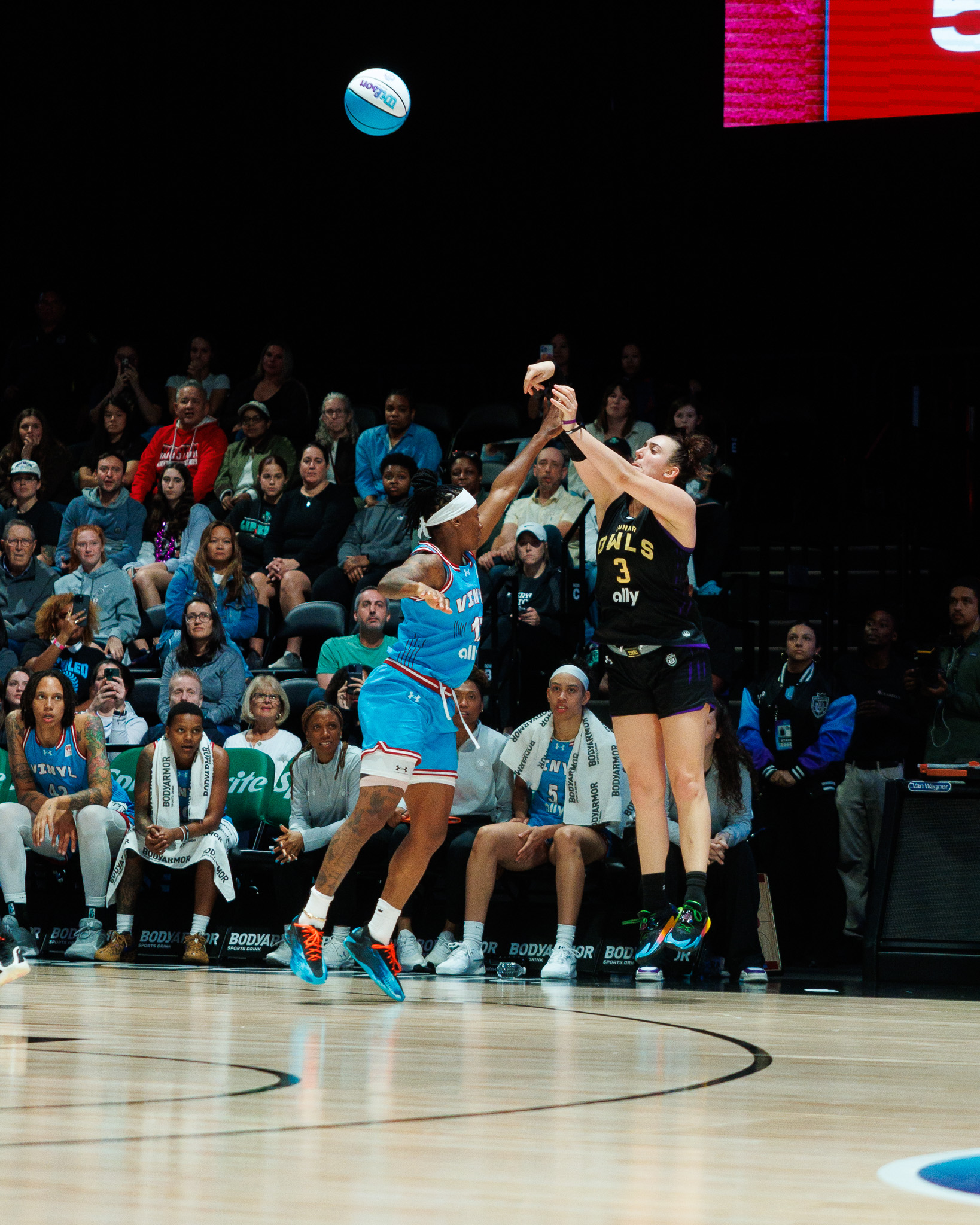 Lunar Owls BC guard Marina Mabrey wearing a black jersey with purple accents jumps to shoot the basketball behind the three point line with ball in the air in front of her. Erica Wheeler is guarding her in a light blue jersey with her hand in Mabrey's face.