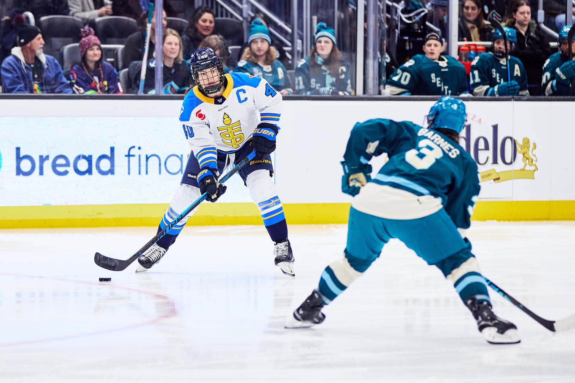 Turnbull looks up ice as she skates with the puck. Cayla Barnes is skating backwards to defend her. Turnbull is in a white away uniform, while Barnes is in a teal home one.