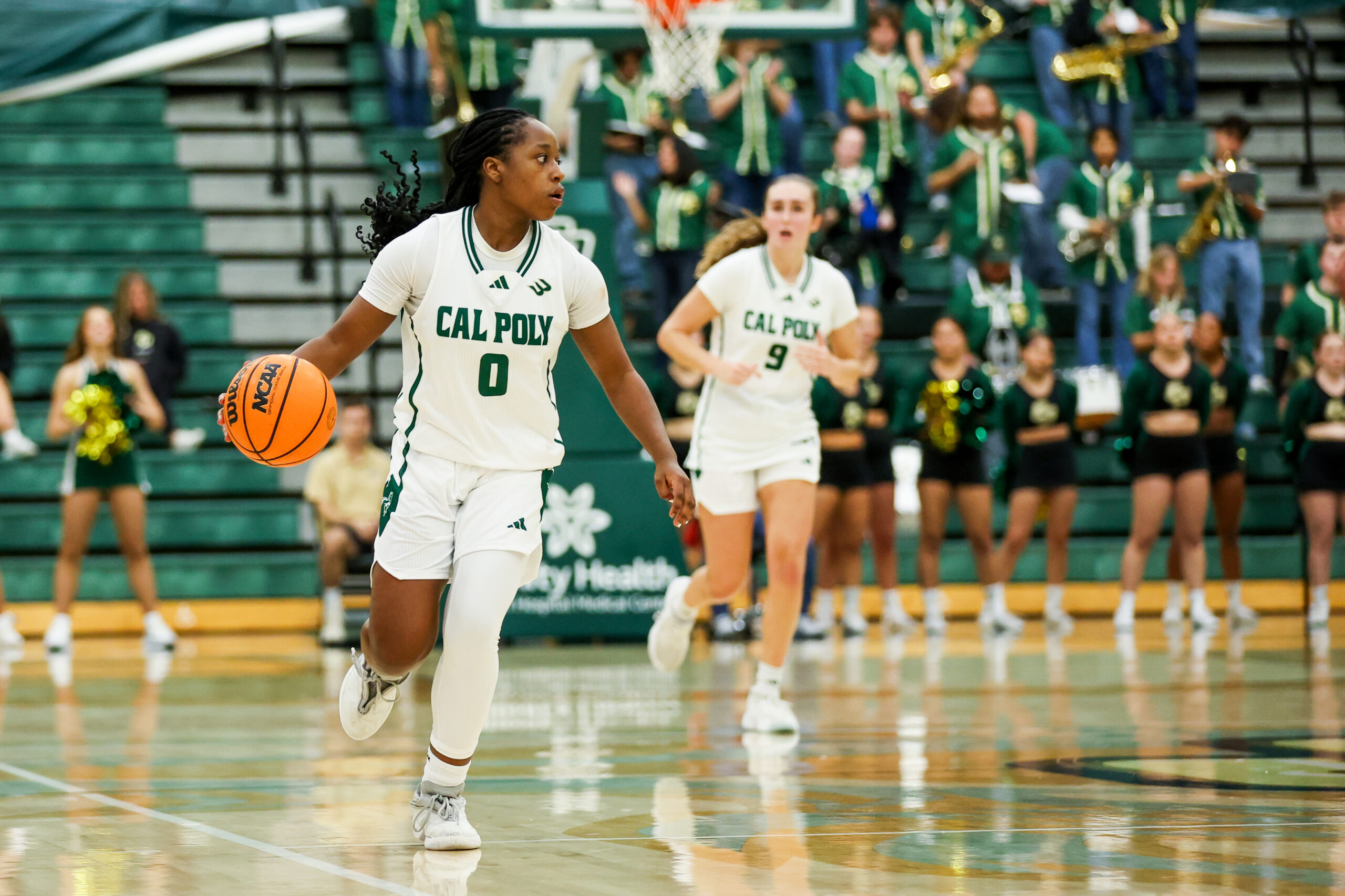 Cal Poly’s Vanessa McManus dribbles the ball up court during a game. The Mustang band and cheer squad are standing behind her.