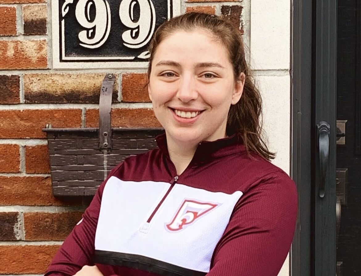 A woman stands in front of a brick building with the number 99 written on it. She is wearing a maroon and white sweater with a stylized letter F on the chest.
