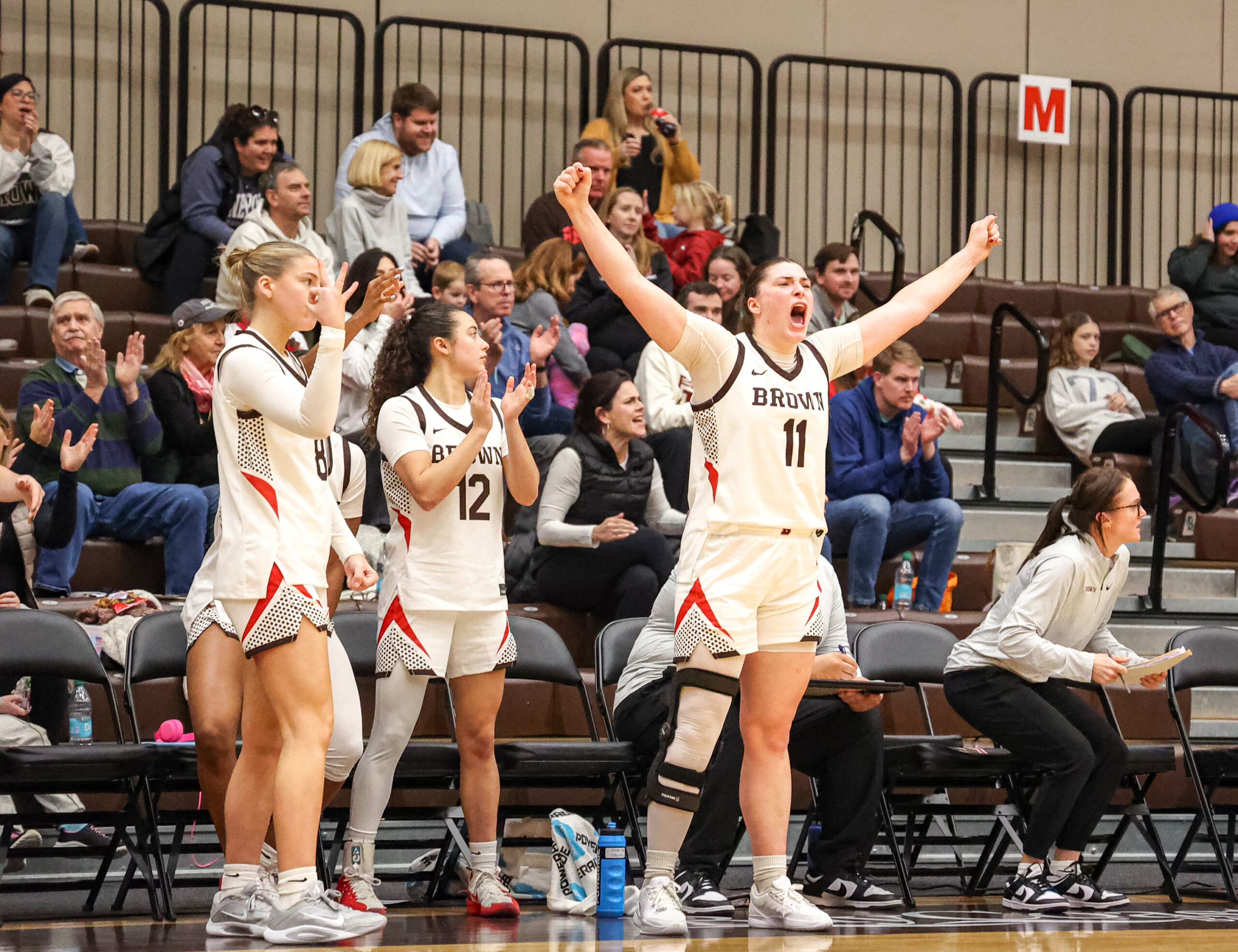 Brown forward Alyssa Moreland stands up on the sideline, extends both arms at 45-degree angles and yells during a game against Penn. Her teammates stand as well, some clapping and some making 3-point signs with their hands.