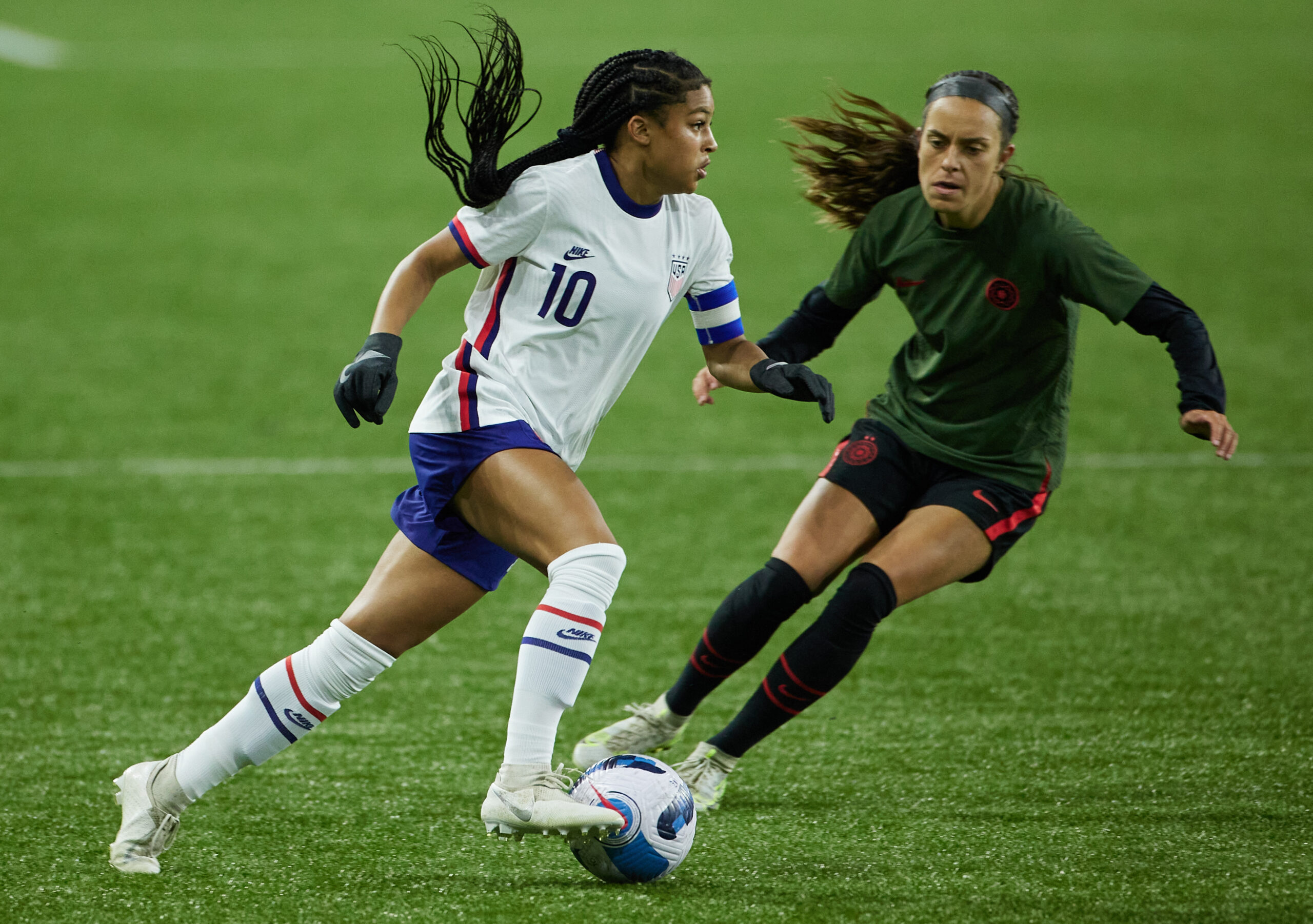A player in a USWNT jersey dribbles the ball, while a player in a dark green jersey challenges her for the ball.