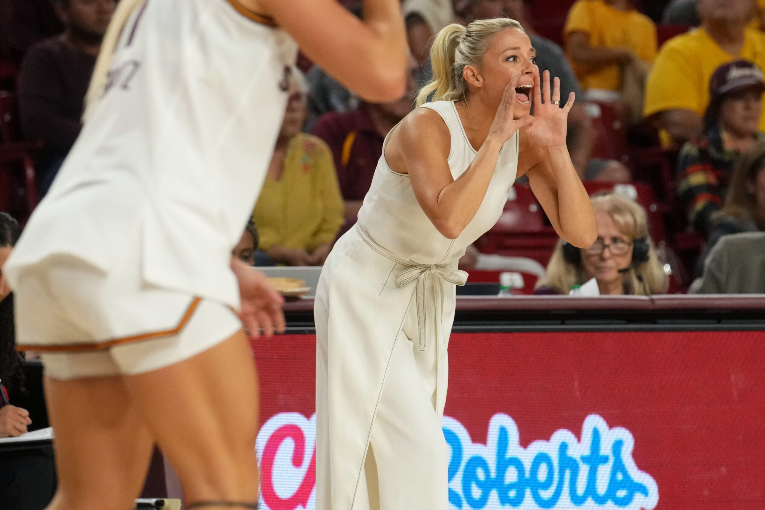 Arizona State women’s basketball head coach Molly Miller, wearing a white sleeveless dress, leans forward on the sideline with her hands cupped around her mouth, shouting instructions to her players during a game. A player in a white uniform is partially visible in the foreground, and fans and staff are seated in the background.