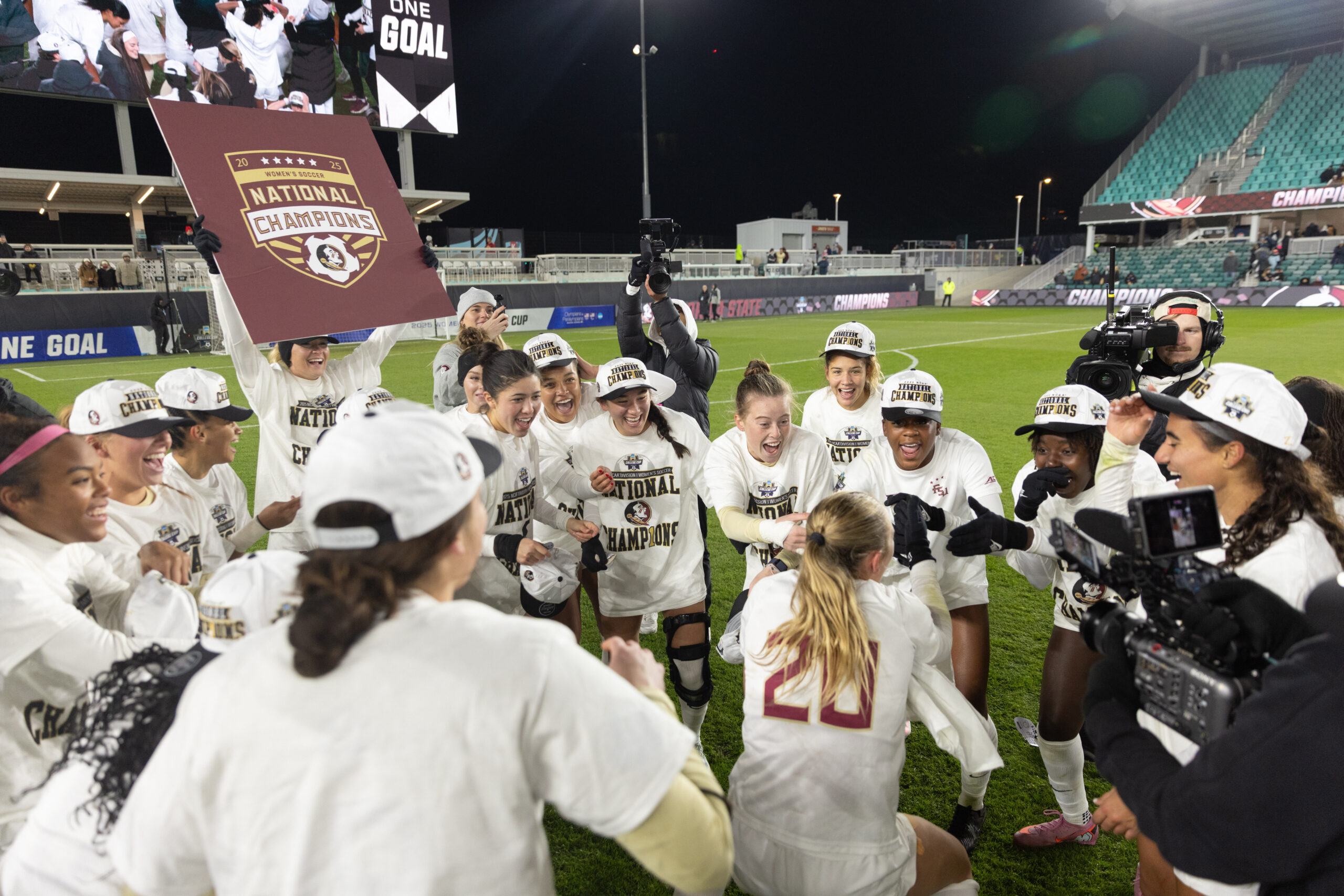 Florida State players standing in a circle and celebrating on the field after the match. One is holding a national championship sign.