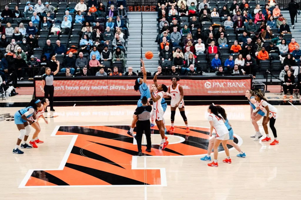 A Columbia player and a Princeton player jump in the air at center court for the opening tip. The eight other starters are standing around the center circle, watching the flight of the ball.