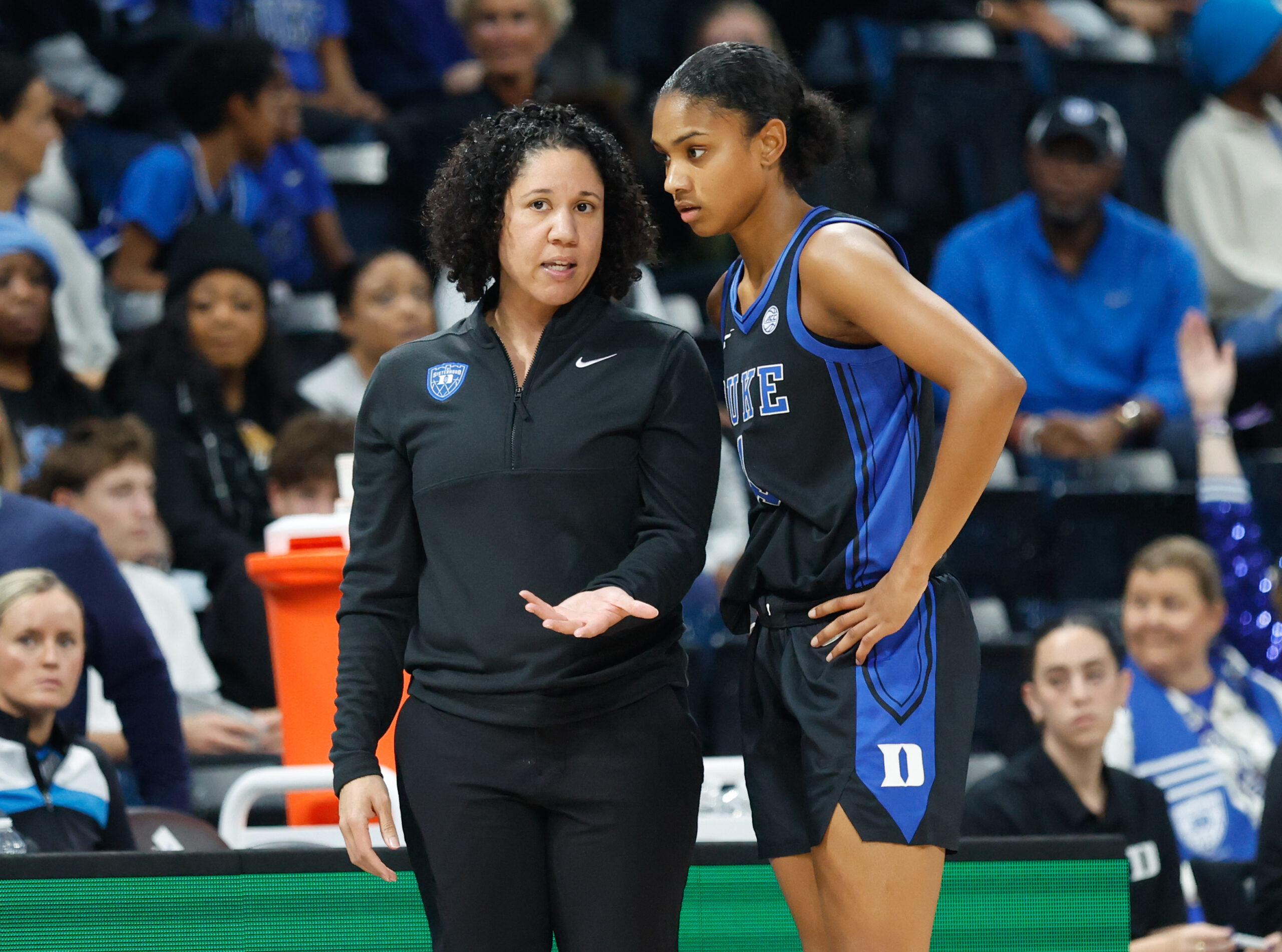 Duke head coach Kara Lawson, wearing a black-quarter zip, offers input from the sideline to guard Riley Nelson, wearing a black and blue Duke jersey.