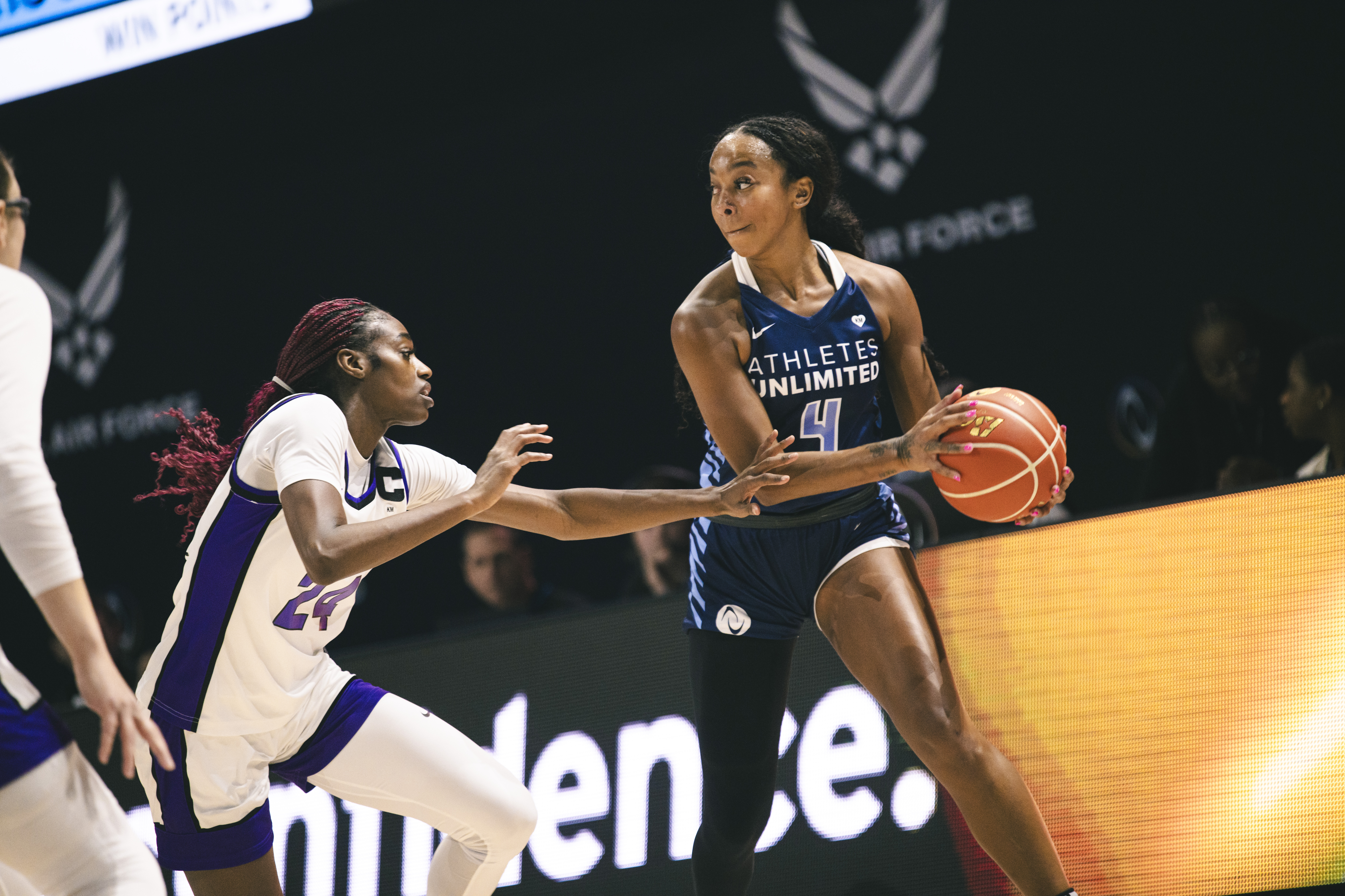 Two players battle for the ball on a basketball court. The player on the left wears a white jersey and the player on the right with the ball wears a dark blue jersey.